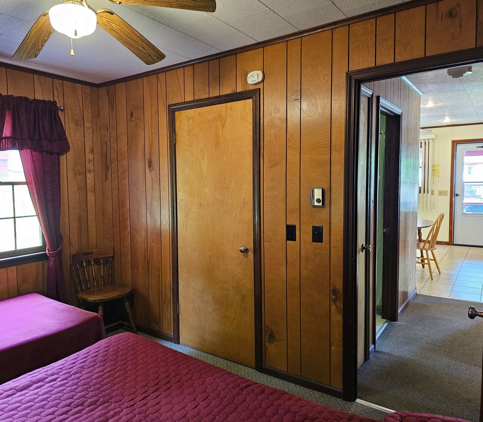 Wood-paneled bedroom with twin beds, ceiling fan, and open doorway to a hallway and dining area.