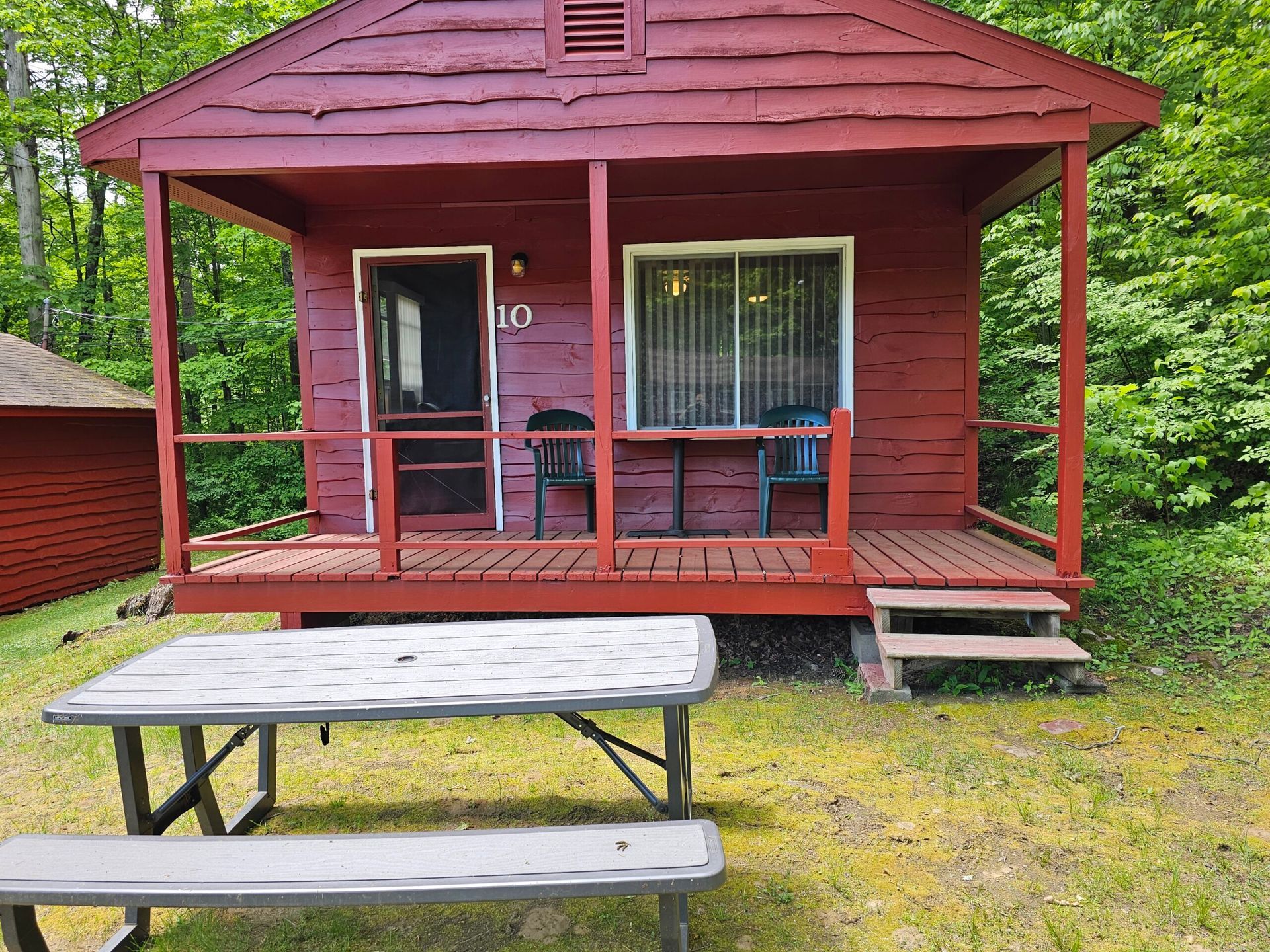 Red cabin porch with two chairs and a picnic table in front, surrounded by green trees.