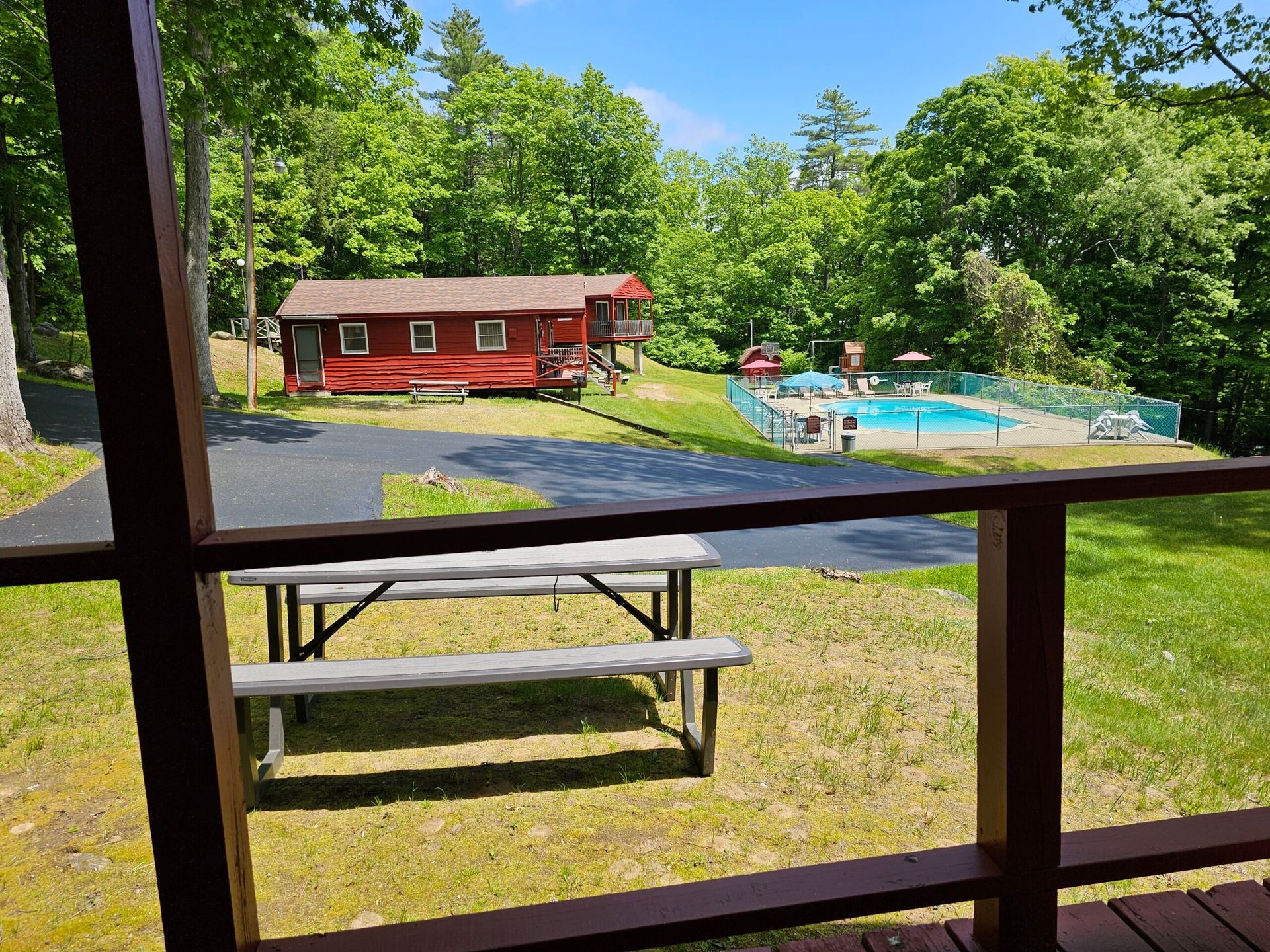 Red cabin by a pool and picnic table, seen through a window on a sunny, wooded day