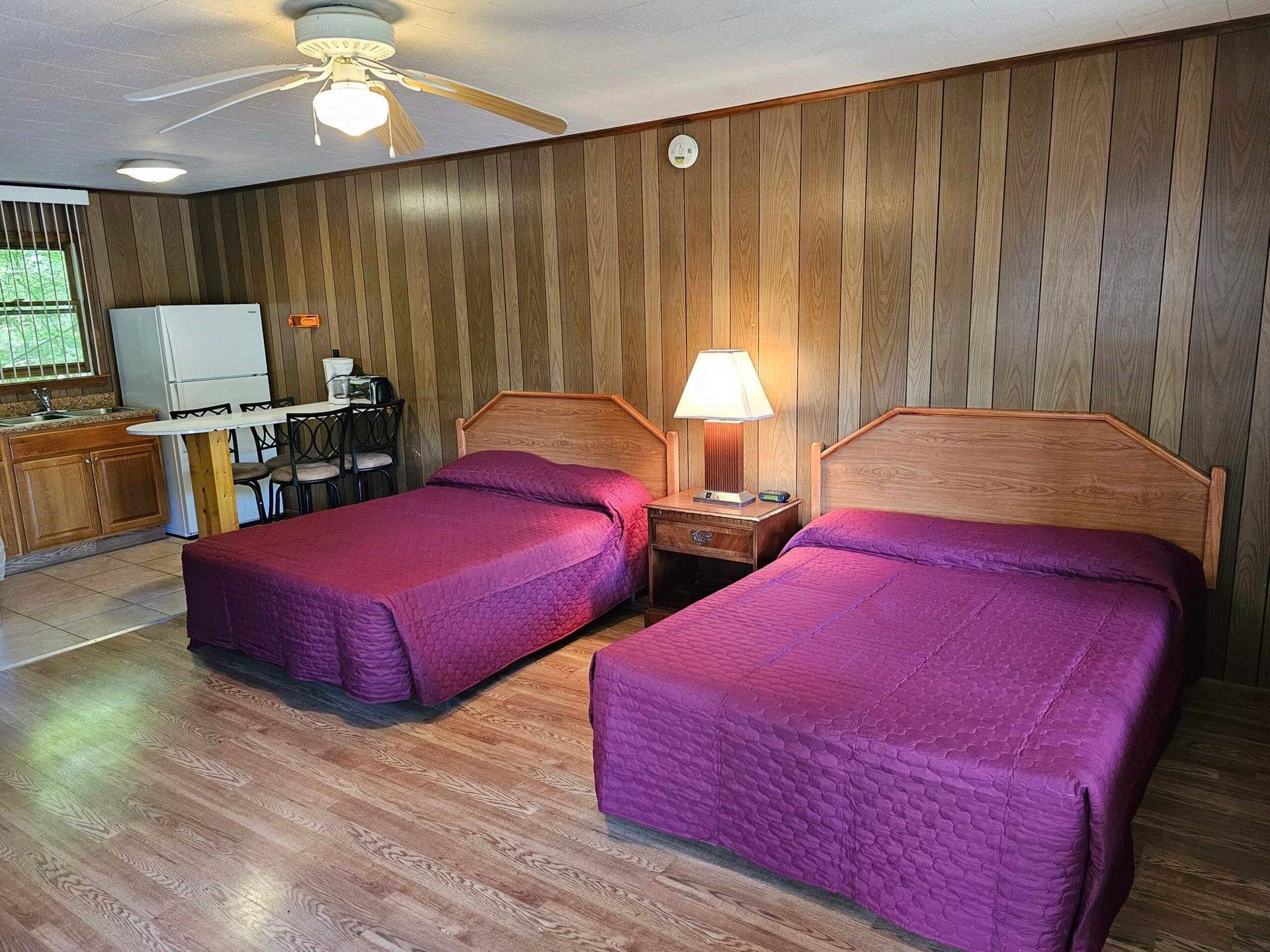 Motel room with two purple-covered beds, wood-paneled walls, nightstand lamp, and kitchenette area.