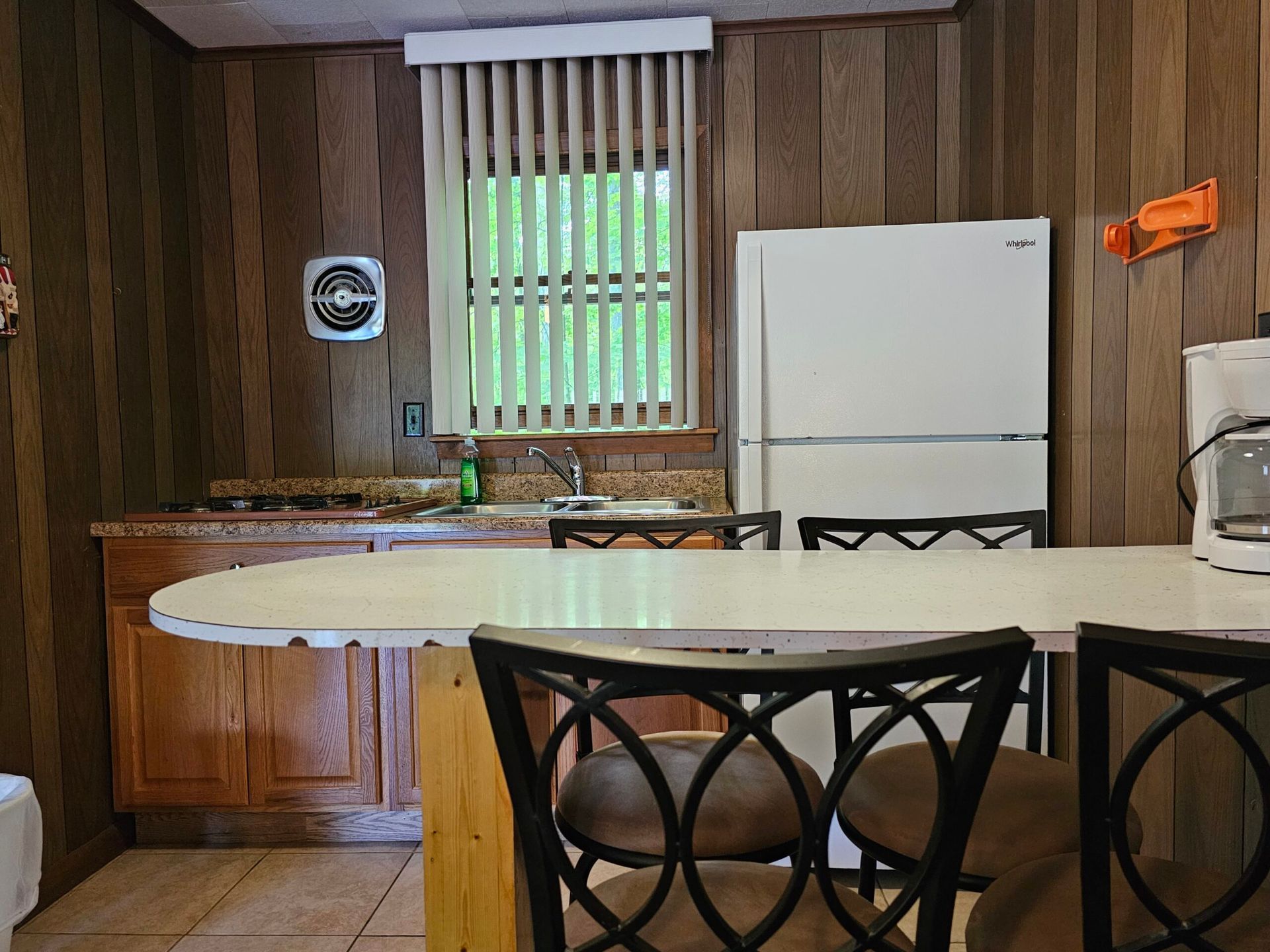 Small kitchen with wood-paneled walls, white fridge, round table, chairs, and a window with vertical blinds.