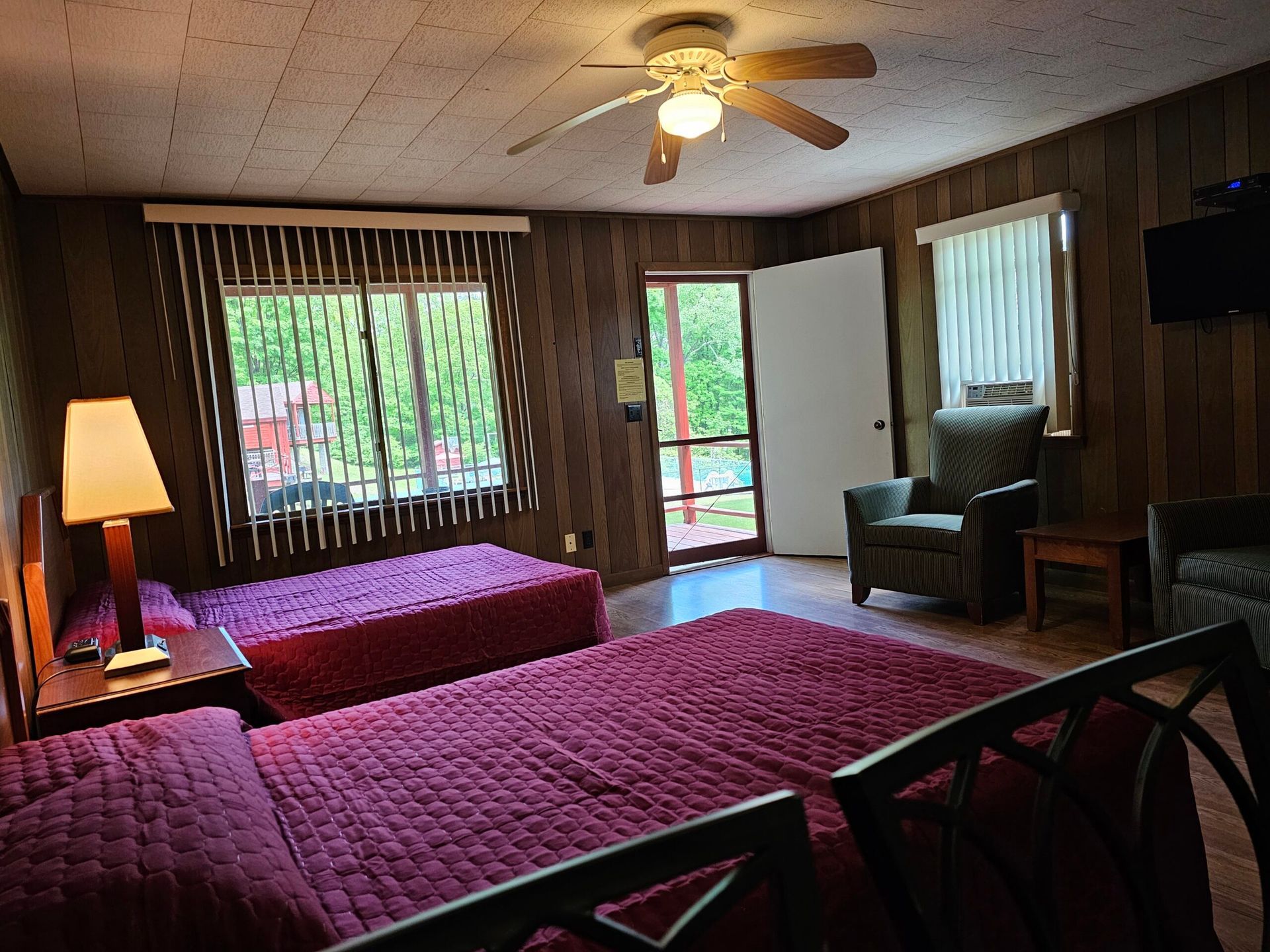 Motel room with two beds, wood-paneled walls, ceiling fan, lamp, chair, and windows with curtains.