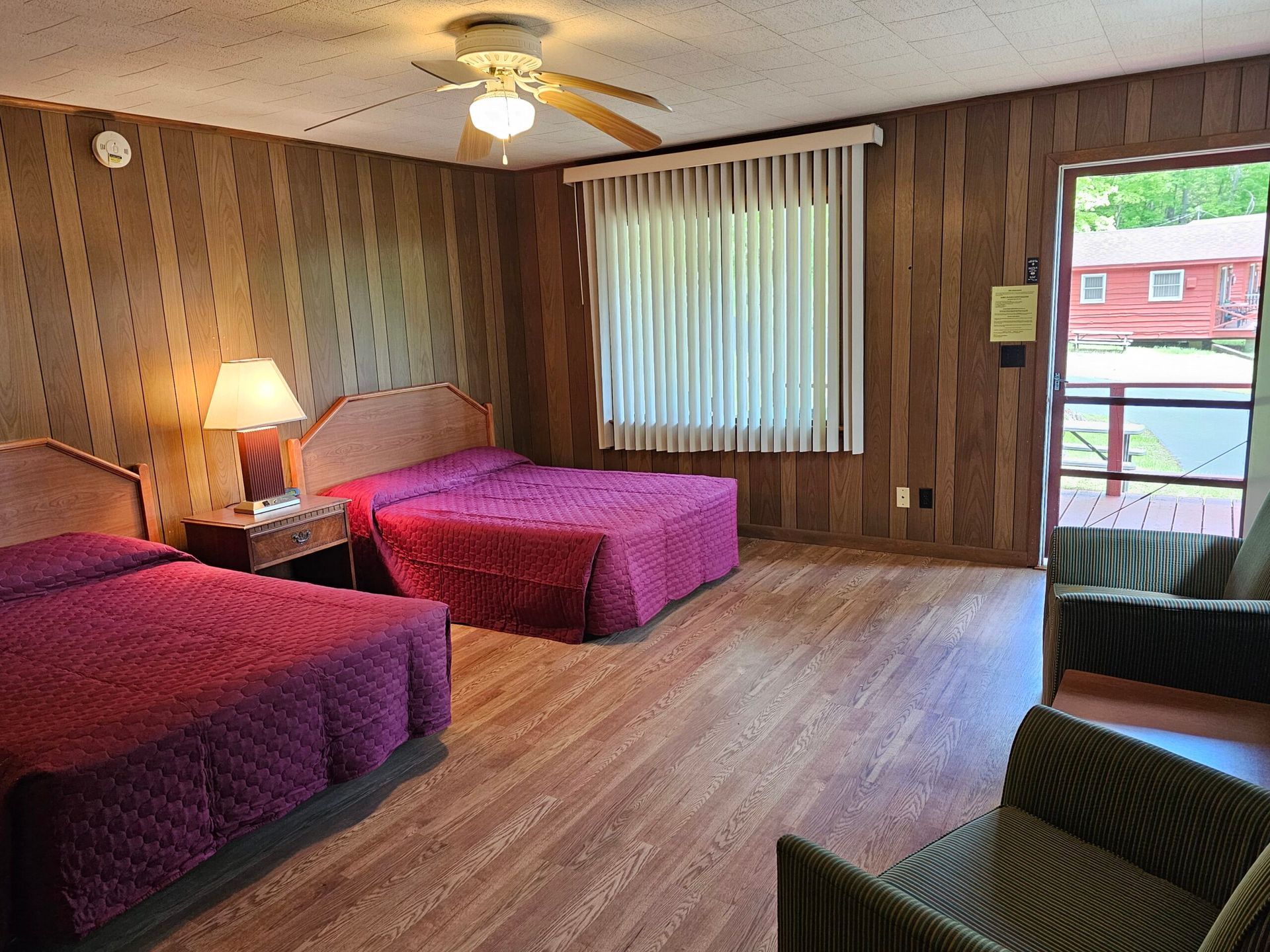 Motel room with two beds, wood-paneled walls, a ceiling fan, and a window with vertical blinds.