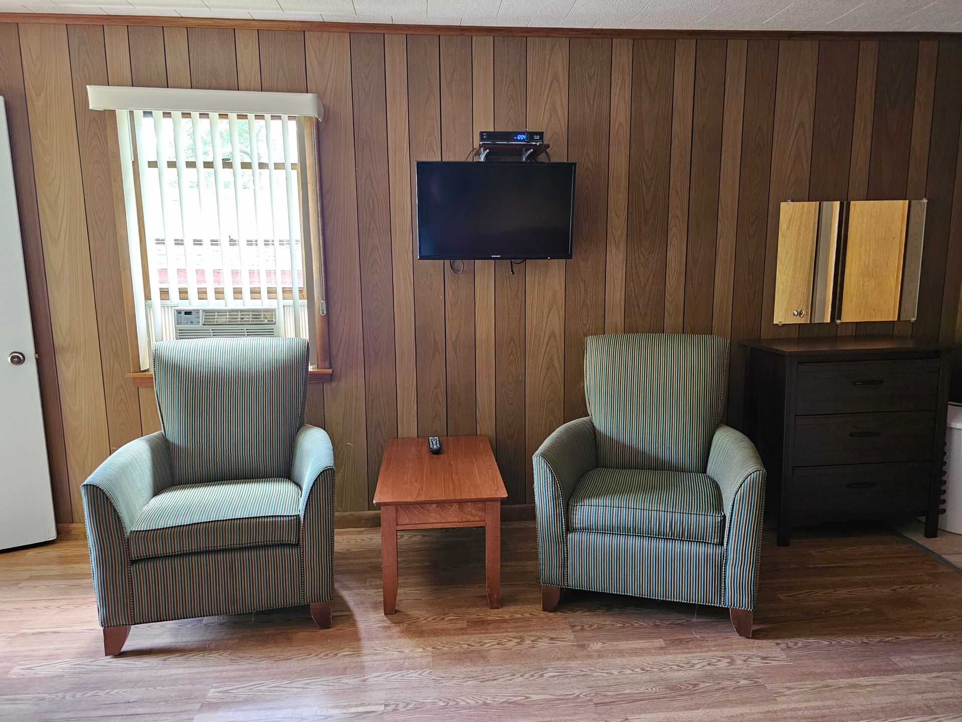 Living room with two striped armchairs, a small table, wall-mounted TV, dresser, and wood-paneled walls.