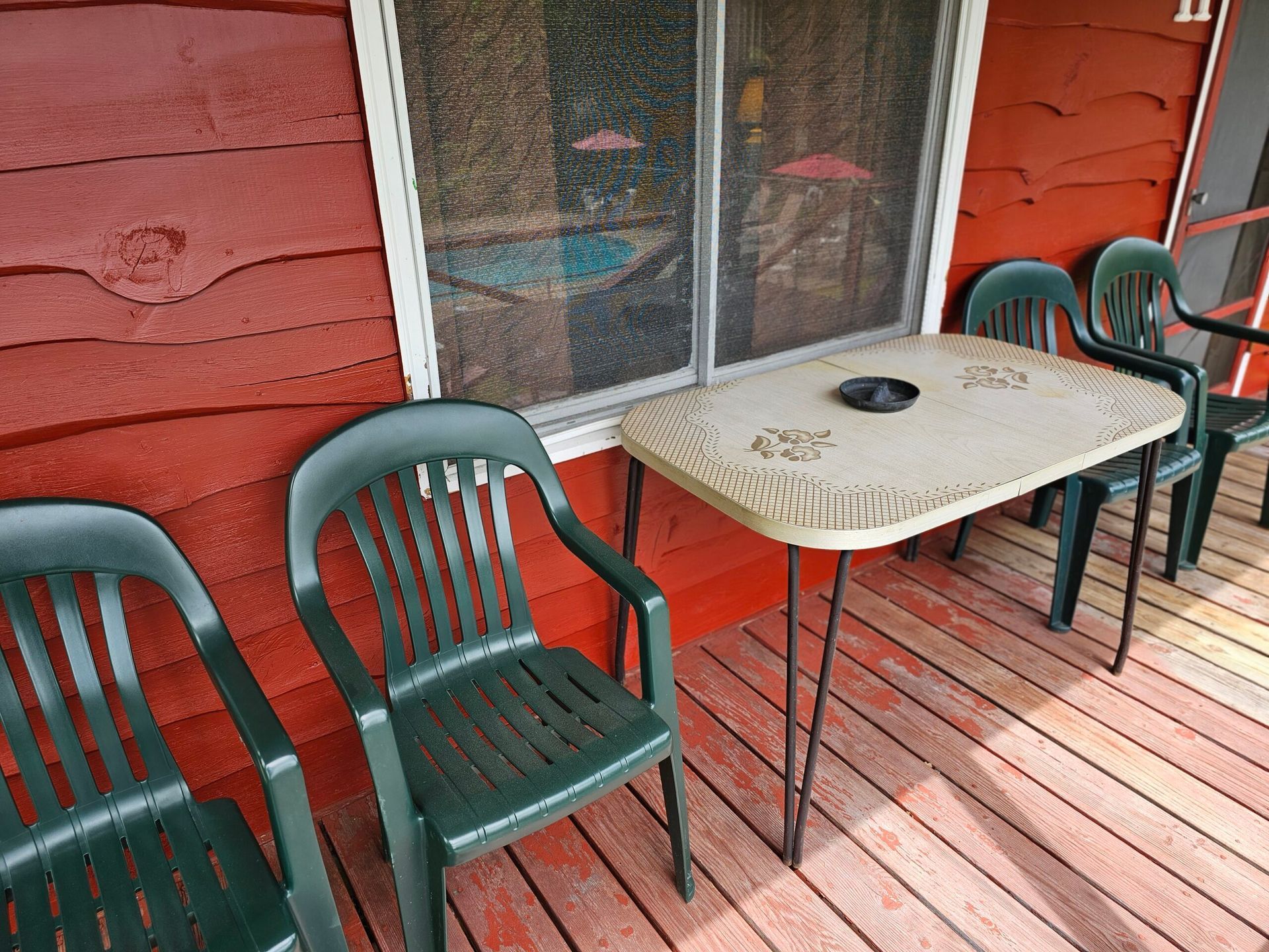 Red porch with three green chairs and a small table beside a screened window.