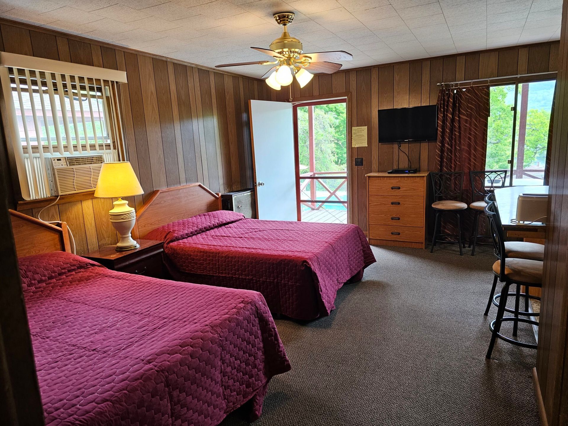Motel room with two red beds, wood-paneled walls, ceiling fan, lamp, TV, and balcony doors to greenery
