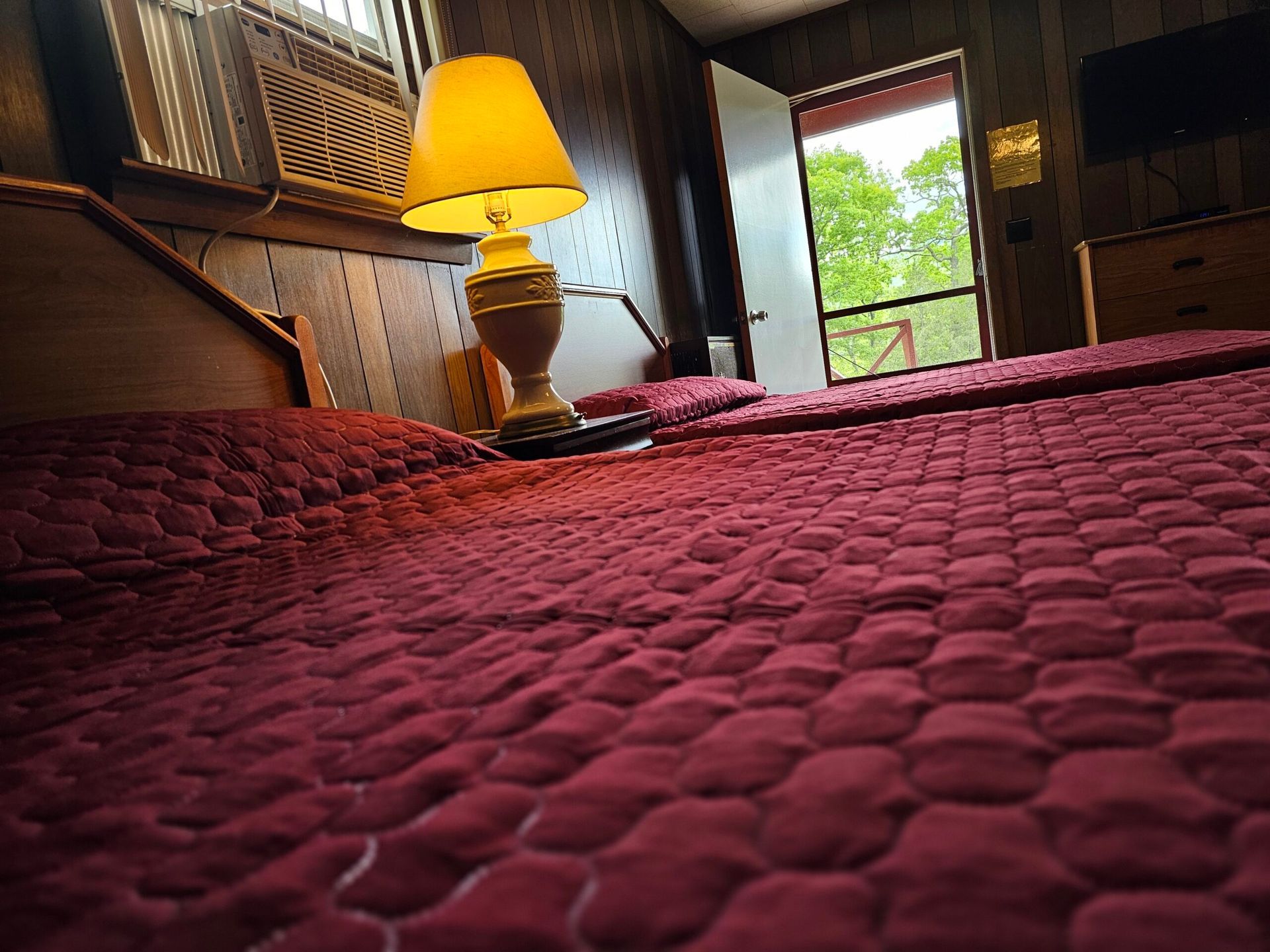 Cozy bedroom with a maroon quilt, lit bedside lamp, and open window showing greenery