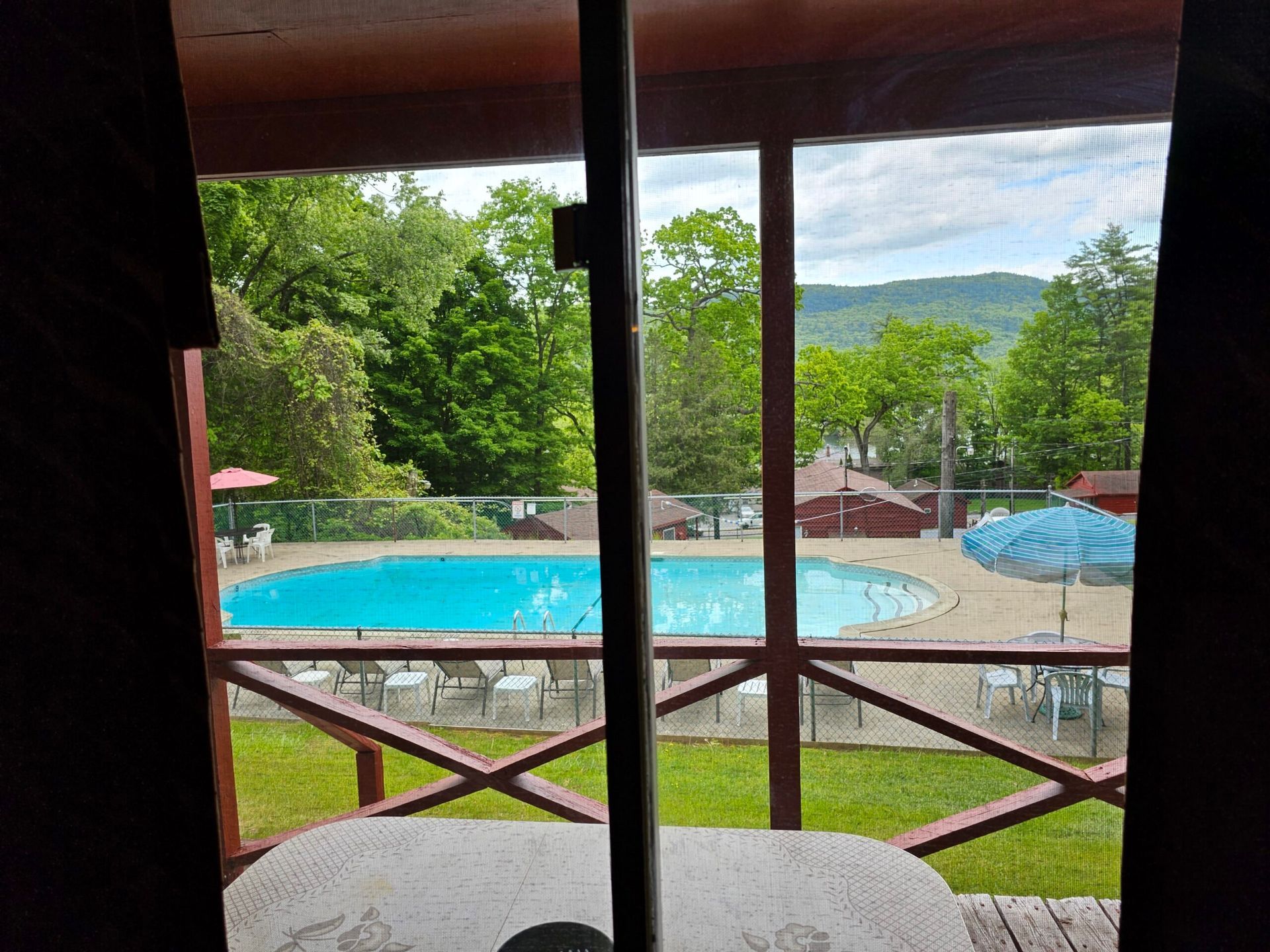 View from a balcony of a turquoise swimming pool, green trees, and distant hills beyond a wooden railing