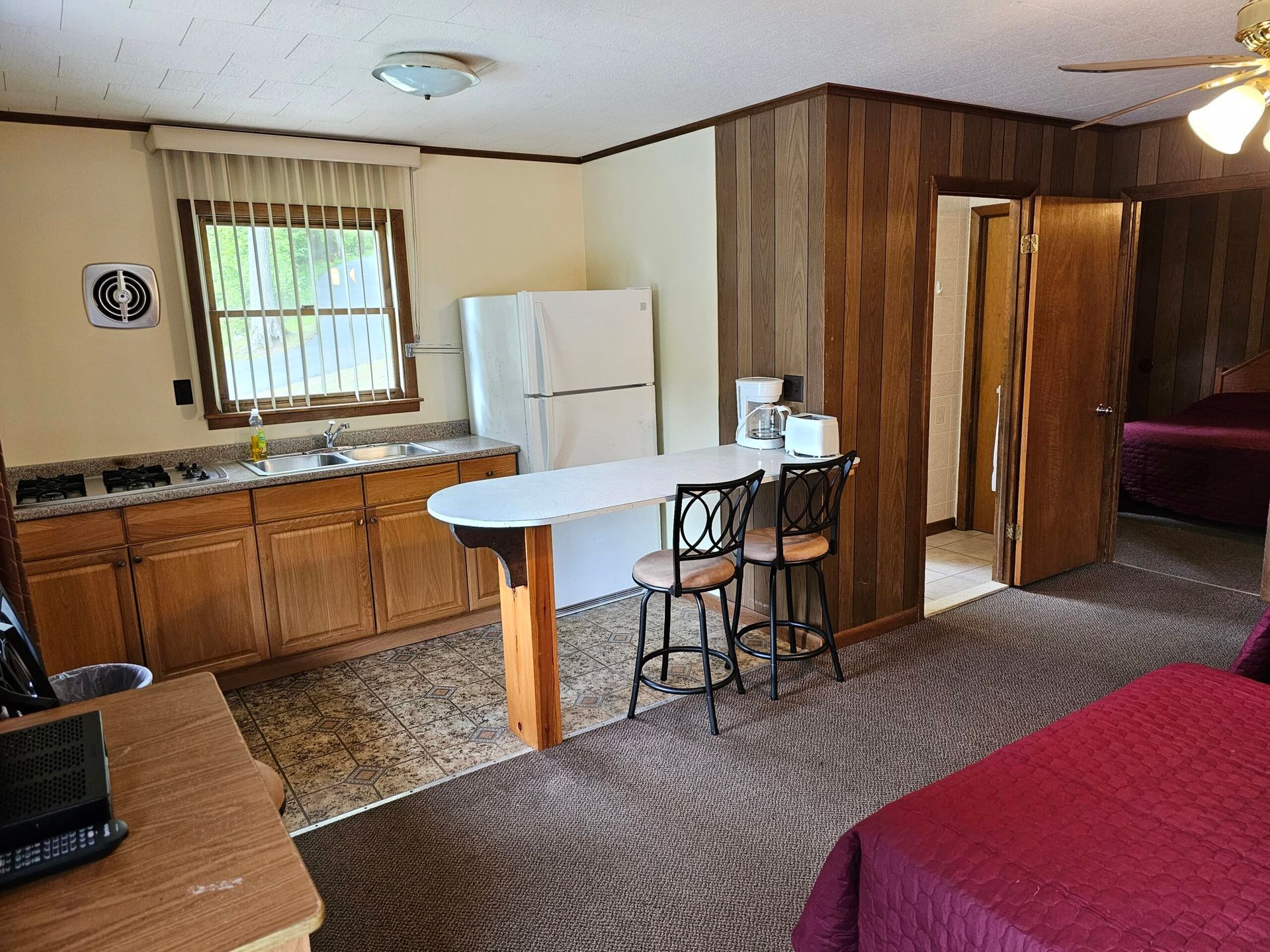Small kitchen with wood cabinets, white fridge, and island; bedroom visible through doorway.