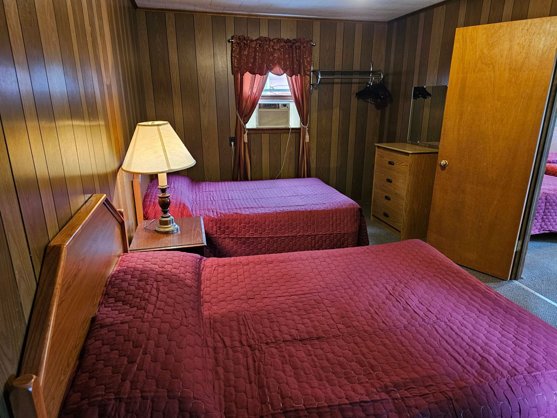 Two twin beds with red quilts in a wood-paneled motel room with a lamp and window.