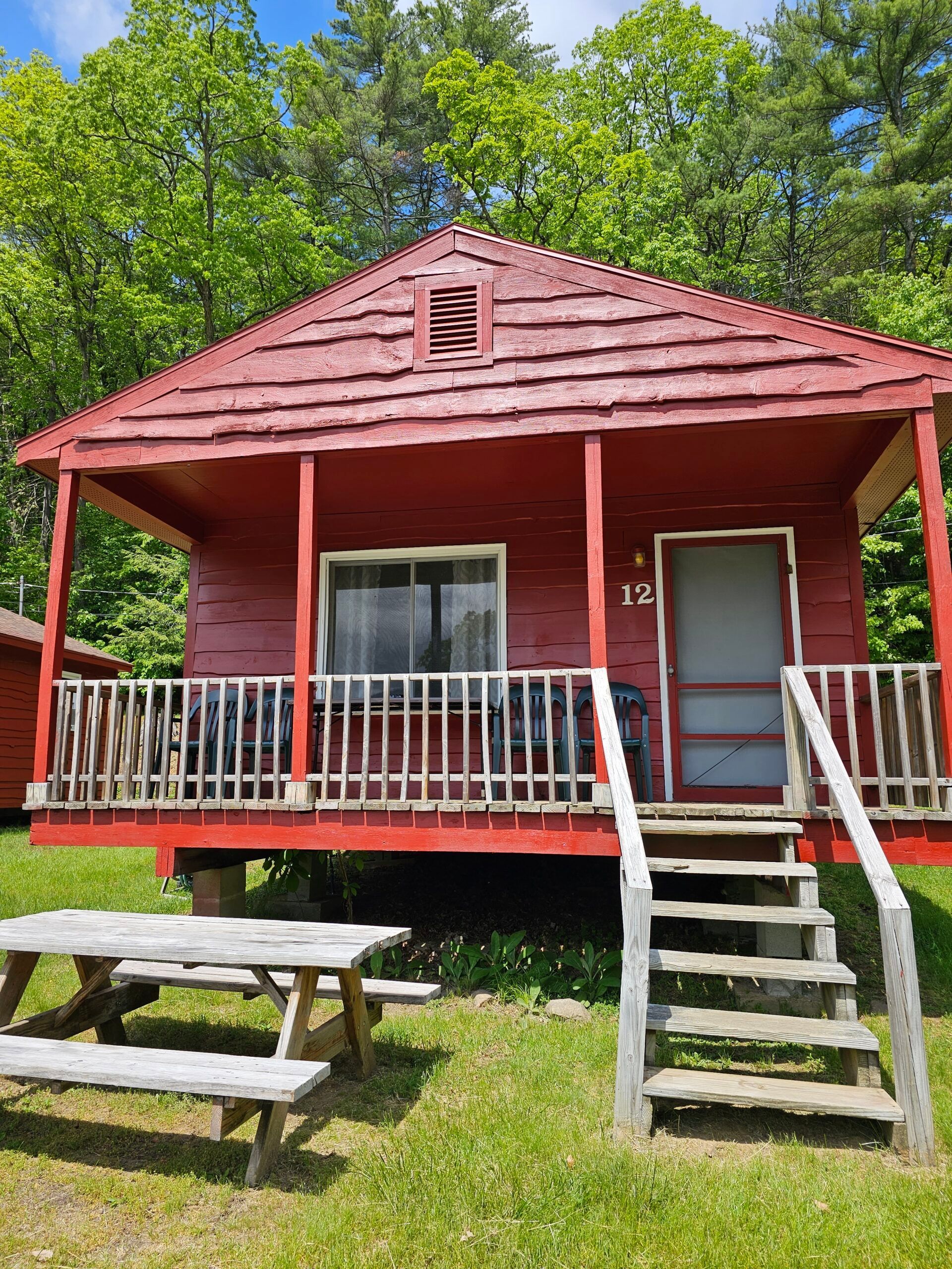 Small red cabin with porch, steps, and picnic table in a grassy wooded area