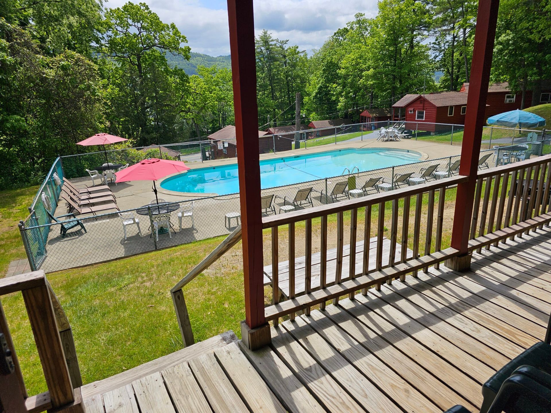View from a porch of a swimming pool with lounge chairs, colorful umbrellas, and trees on a sunny day