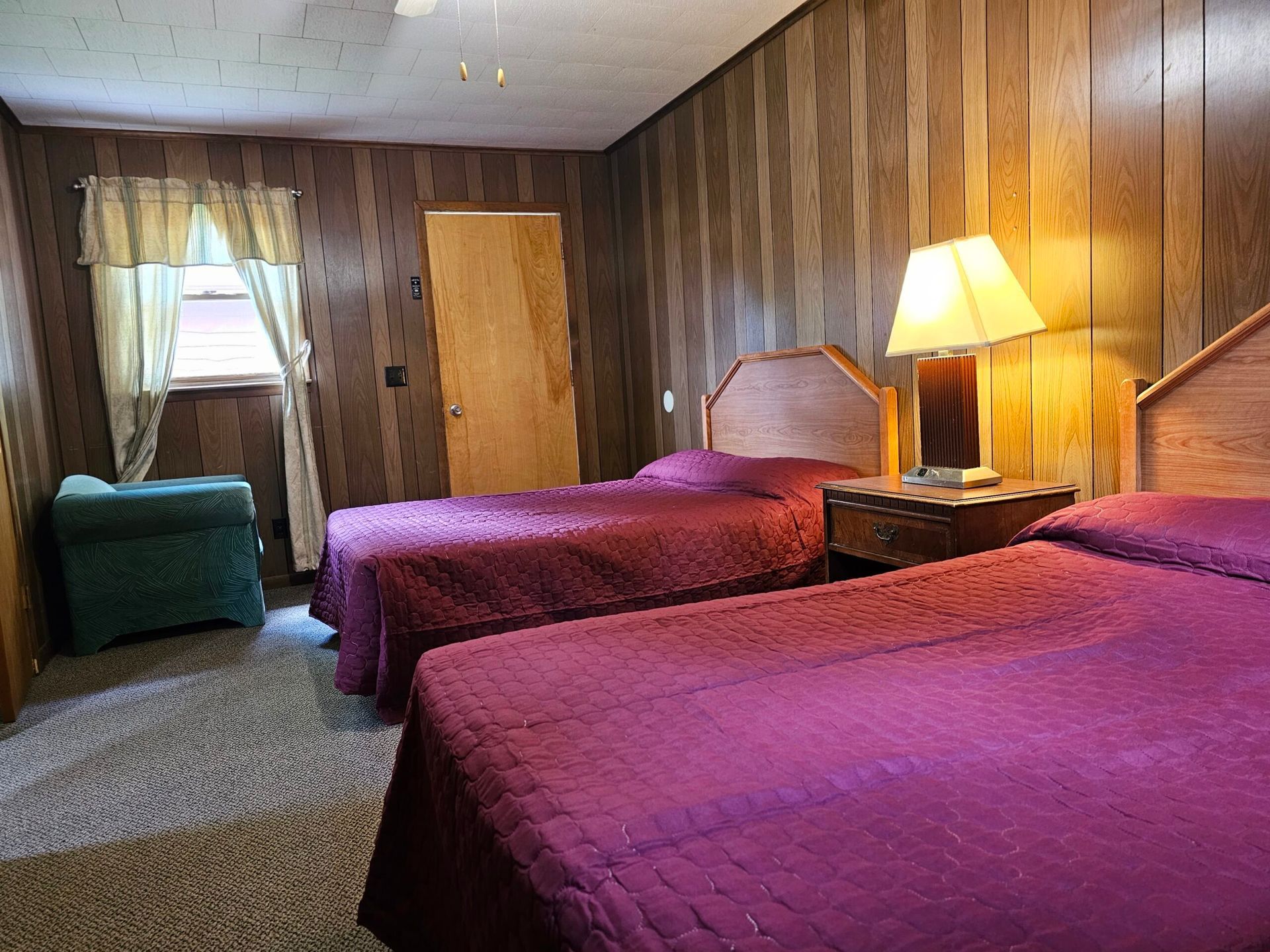 Motel room with two purple beds, wood-paneled walls, lamp, and window with curtains