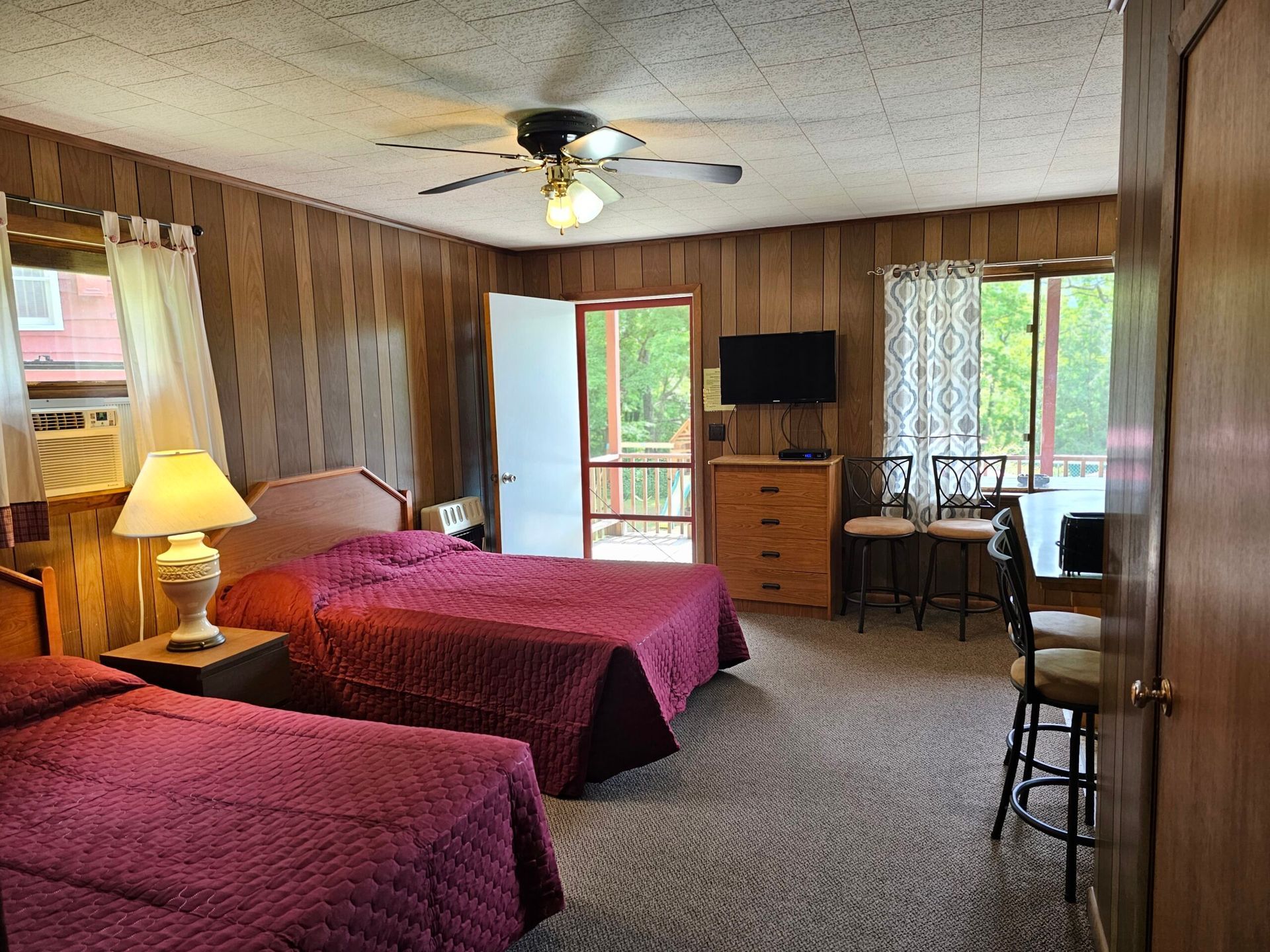 Bedroom with two red beds, wood-paneled walls, ceiling fan, TV, and sliding glass door to a balcony.