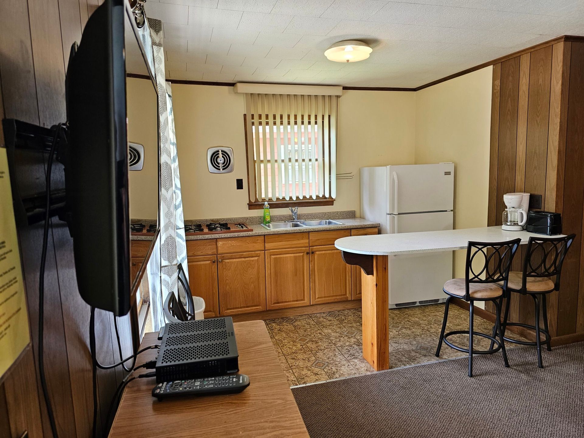 Small kitchen with wood cabinets, white refrigerator, breakfast table, TV, and dark wood paneling.