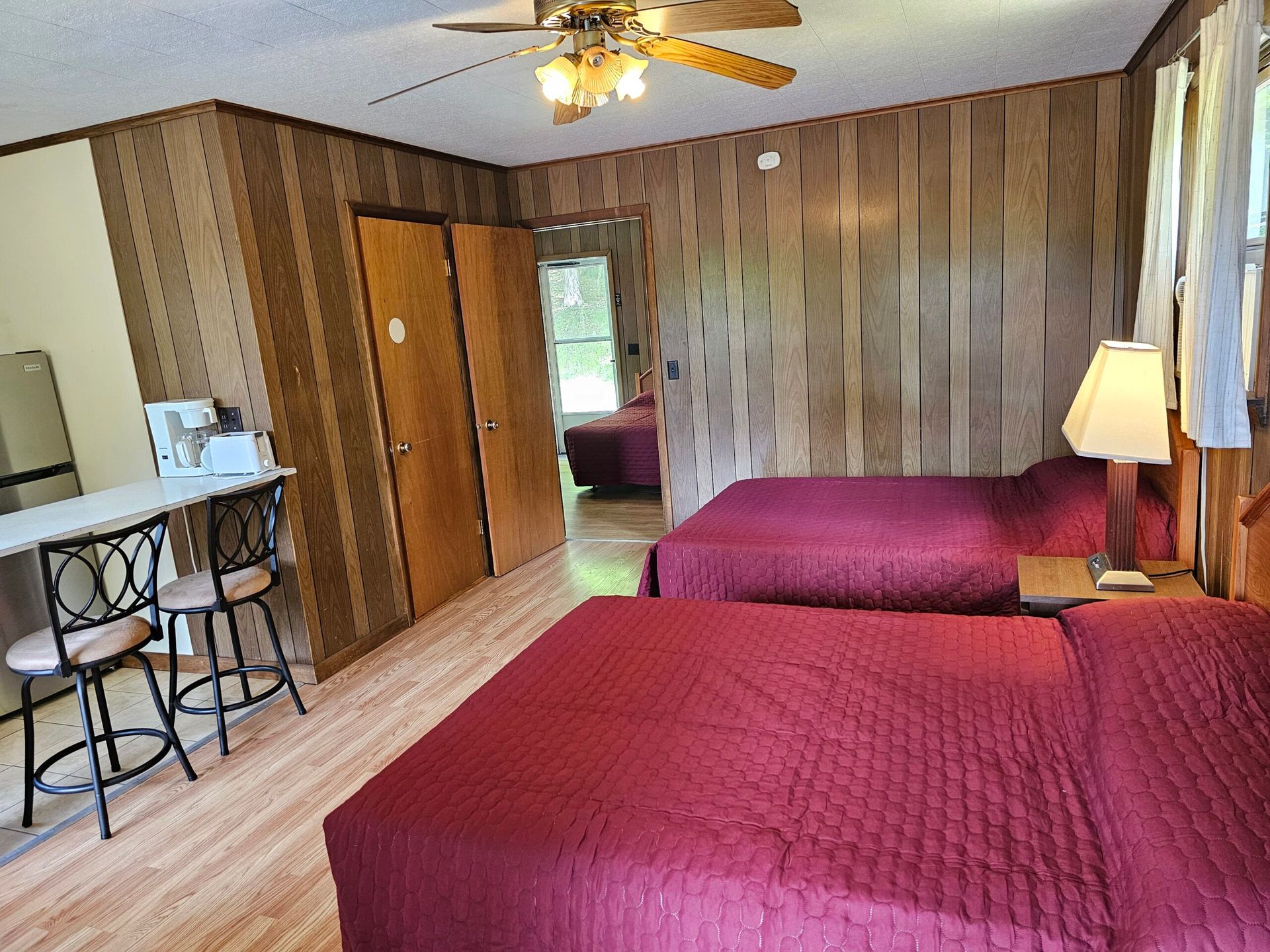 Bedroom with two red beds, wood-paneled walls, ceiling fan, and a small table by the window.