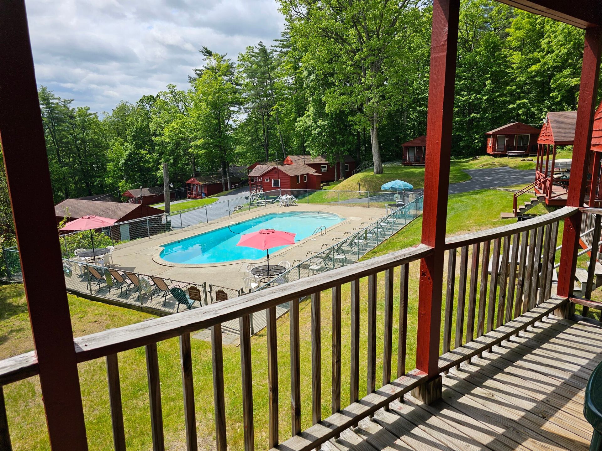 Outdoor resort pool with pink umbrellas, surrounded by trees and cabins, viewed from a wooden balcony.