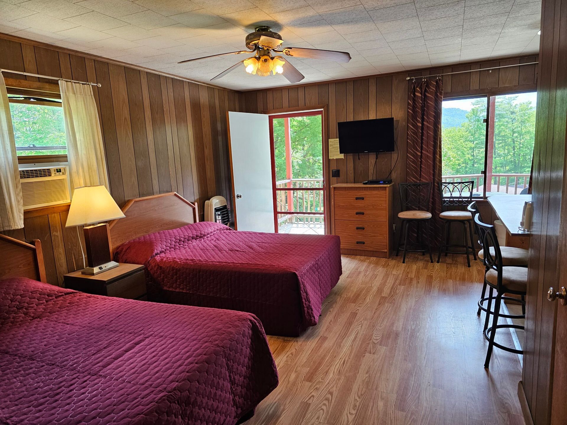Motel room with two red beds, wood-paneled walls, ceiling fan, TV, desk, and balcony door.