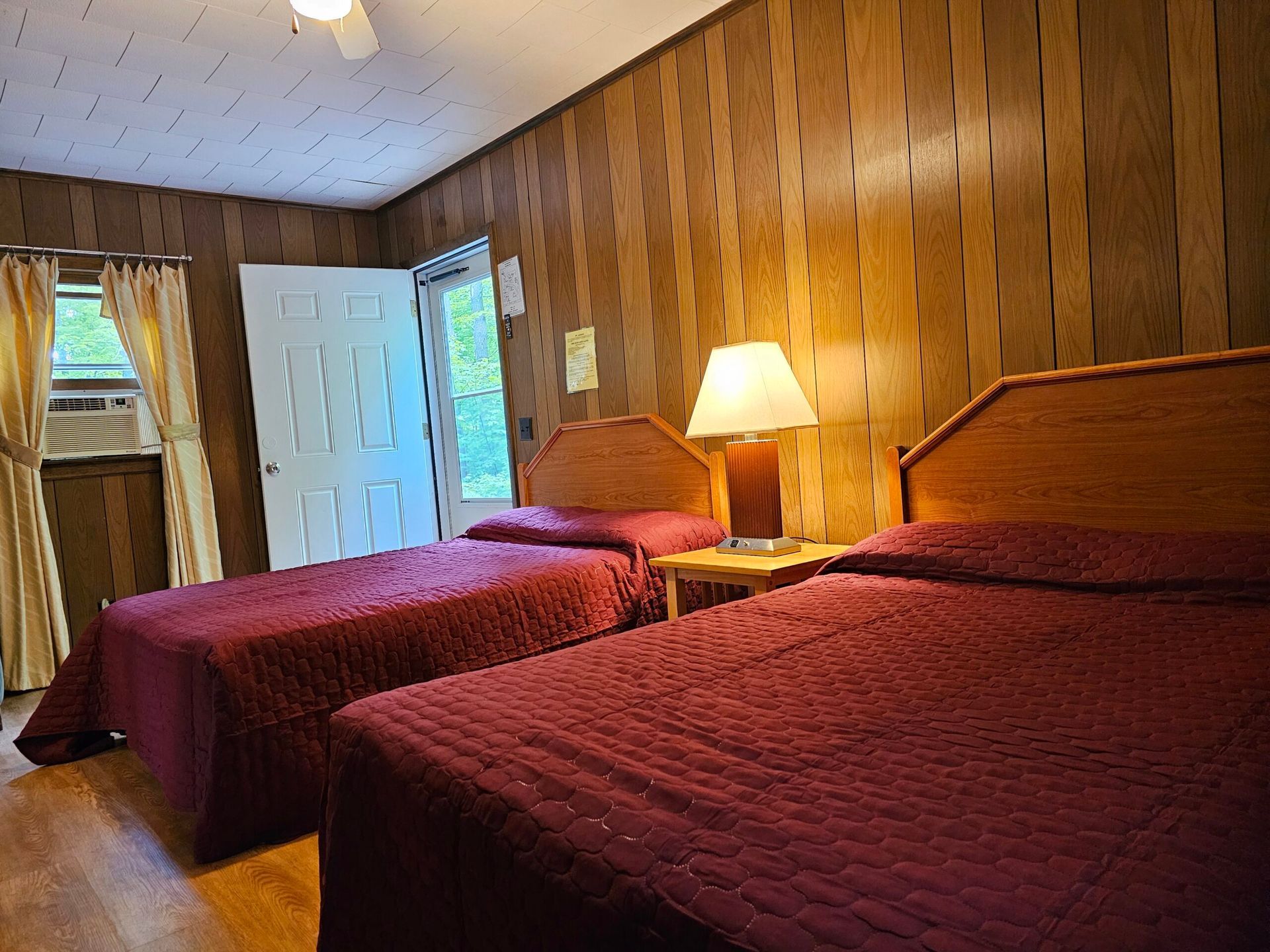 Wood-paneled bedroom with two twin beds, red quilts, and a bedside lamp near an open door.