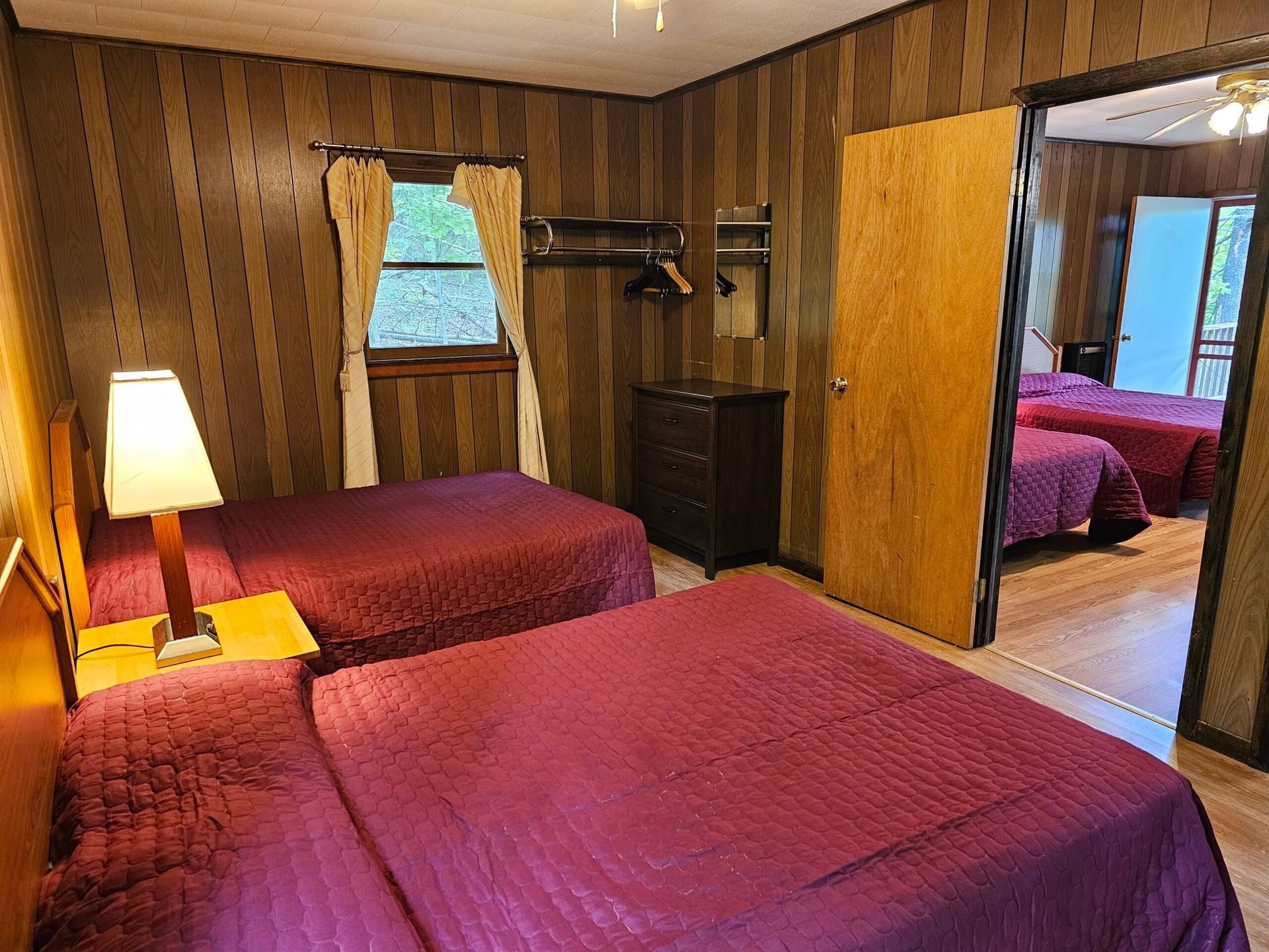 Bedroom with two red beds, a lit lamp, wood-paneled walls, and an open doorway to a second room
