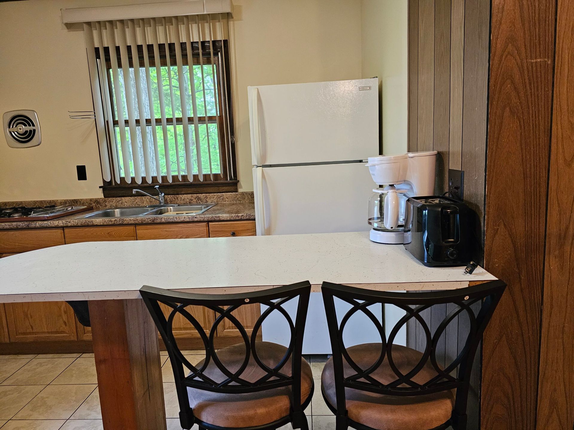 Small kitchen with white refrigerator, window, countertop appliances, and two black chairs at a white table.