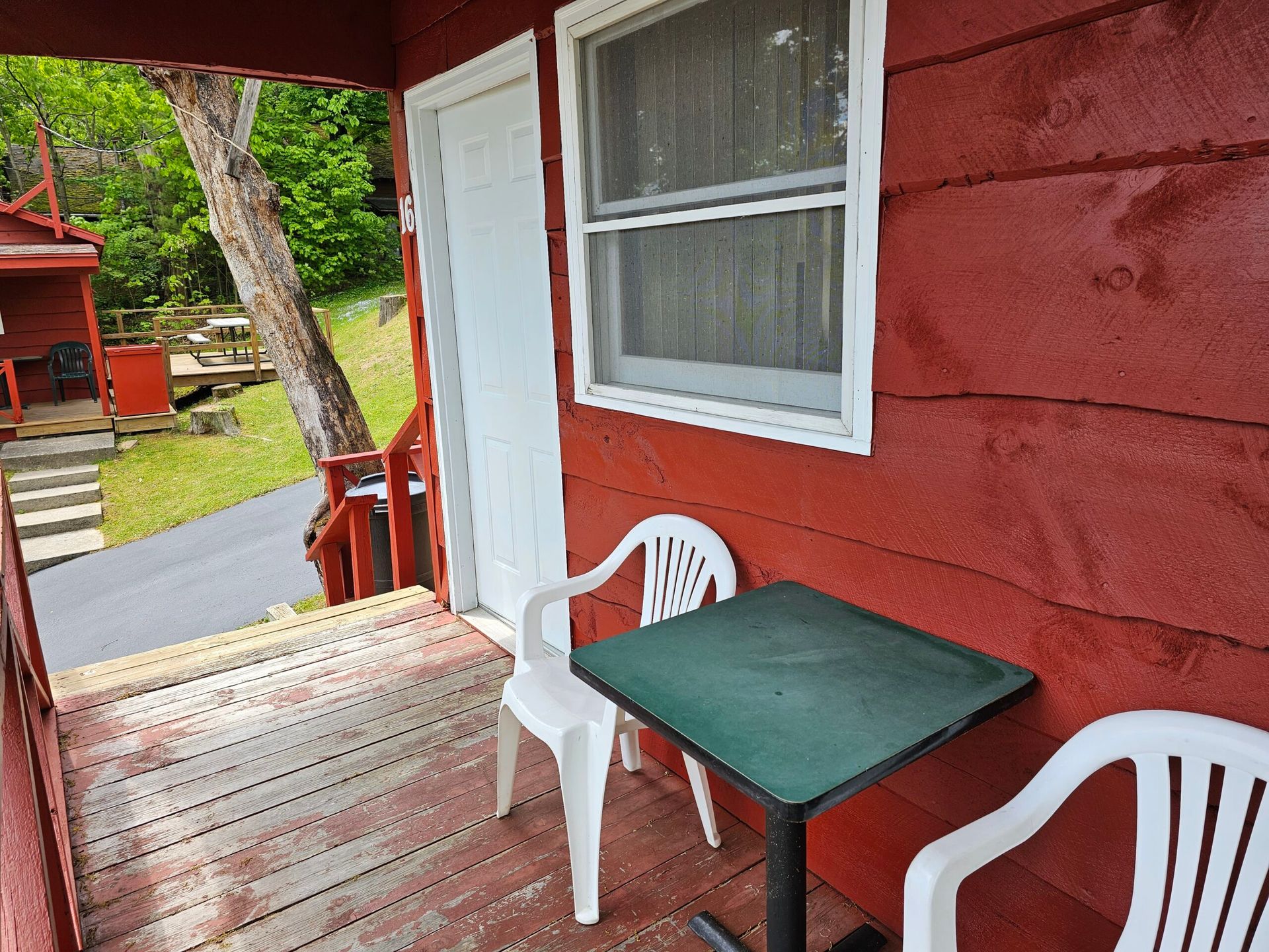 Small porch with red siding, white door and window, green table, and white chairs by a driveway