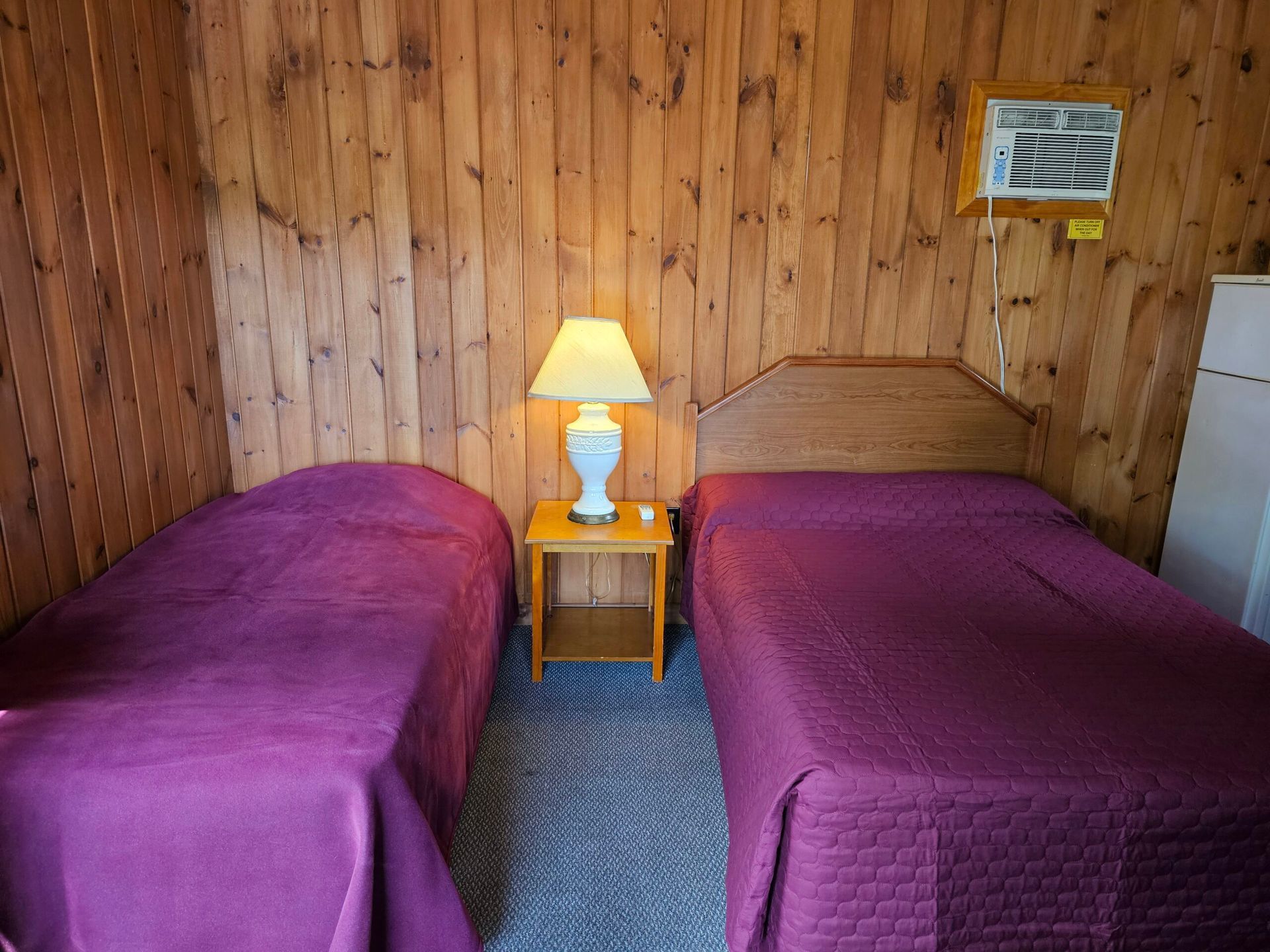 Two beds with purple bedspreads in a wood-paneled room, with a lamp on a nightstand between them.