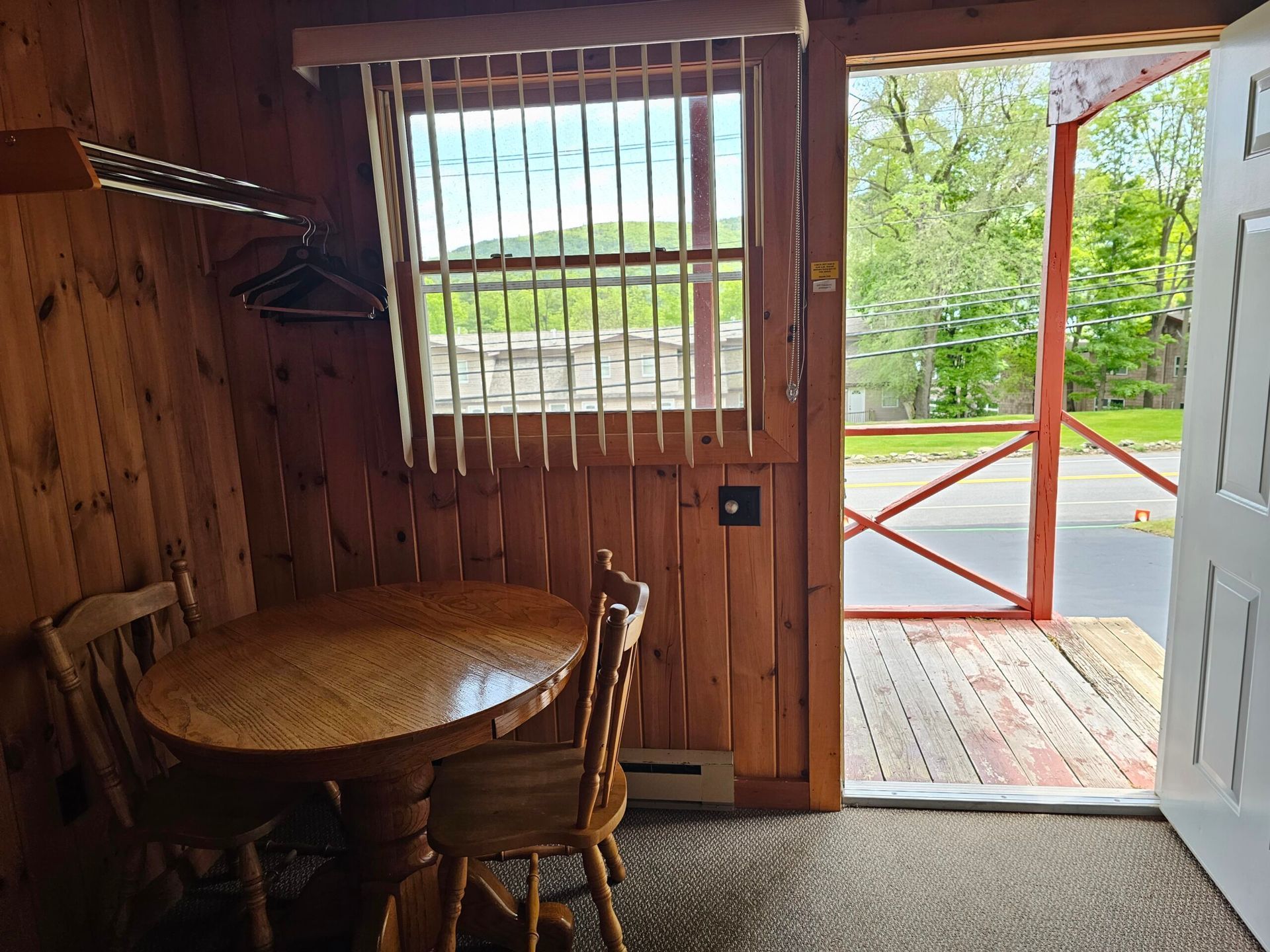 Wooden cabin interior with round table and chair, open door to a sunlit porch and trees outside