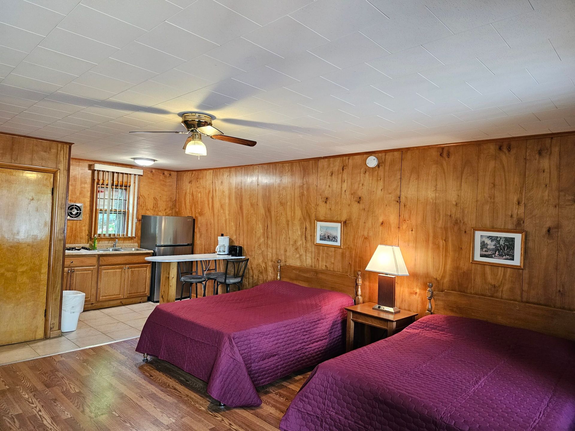Cozy wood-paneled bedroom with two purple beds, small table, lamp, and kitchenette area
