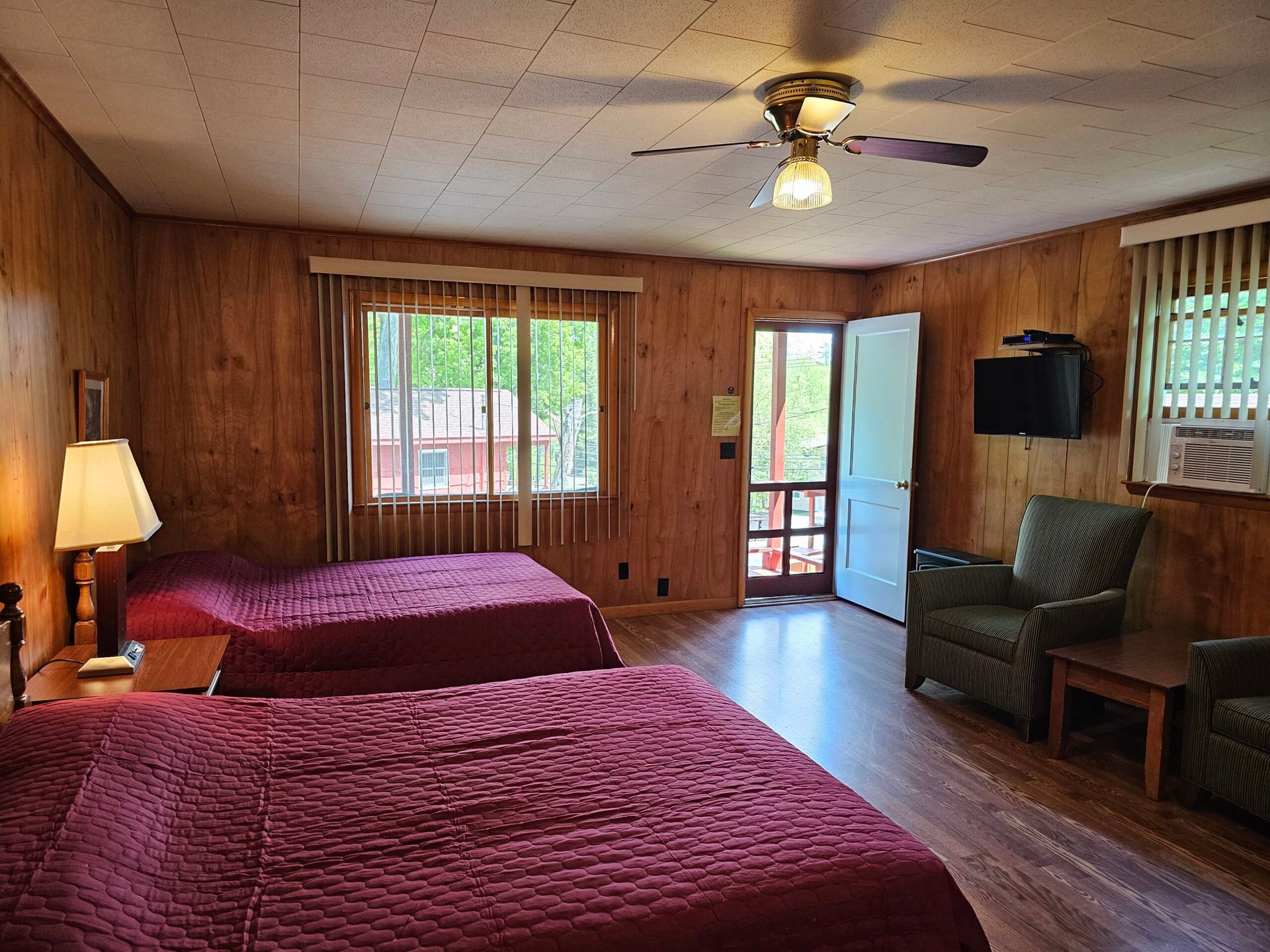 Wood-paneled motel room with two beds, a lamp, chairs, and windows letting in daylight.