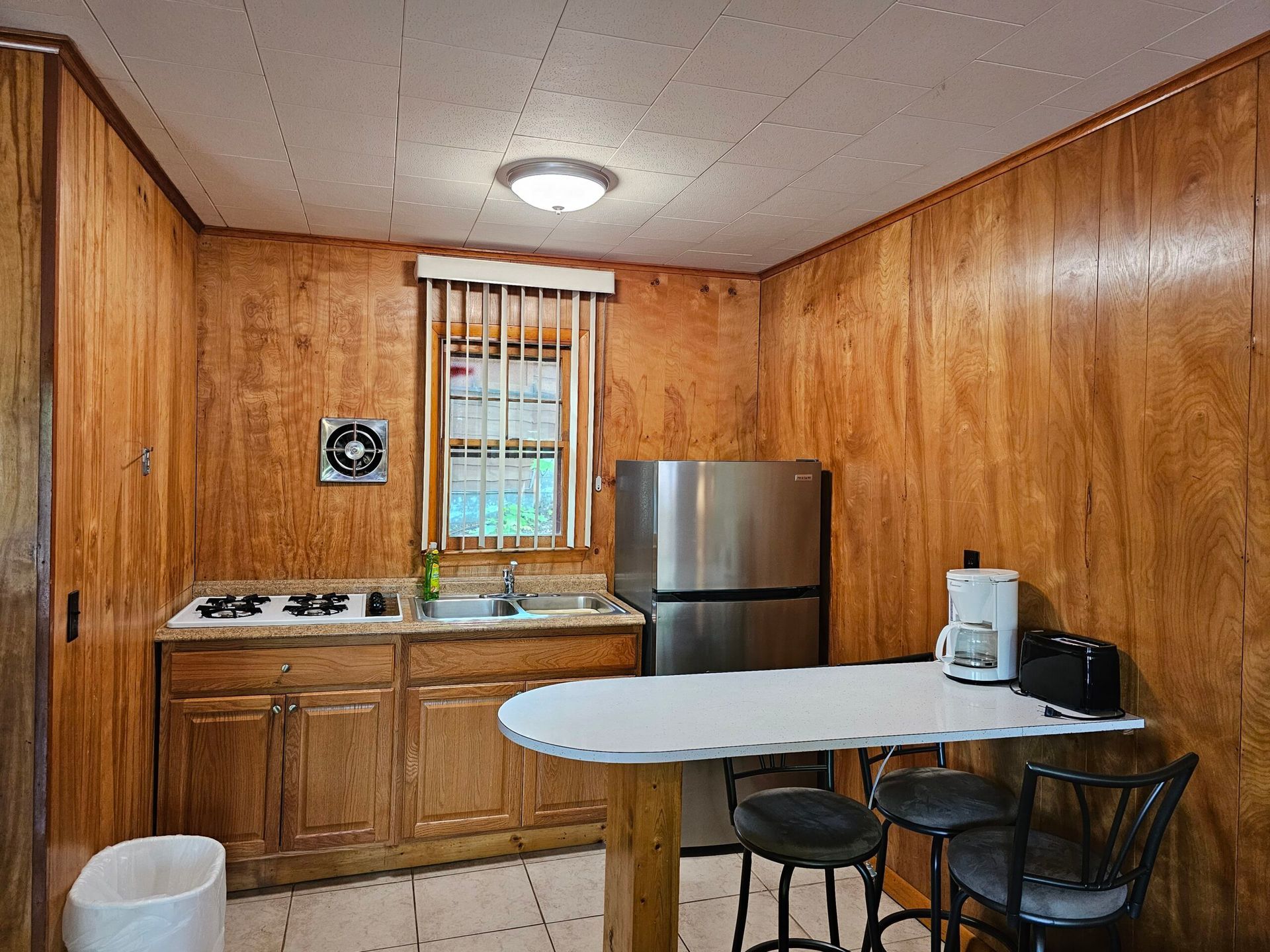 Small kitchen with wood-paneled walls, sink, fridge, oval table, and two chairs under a ceiling light