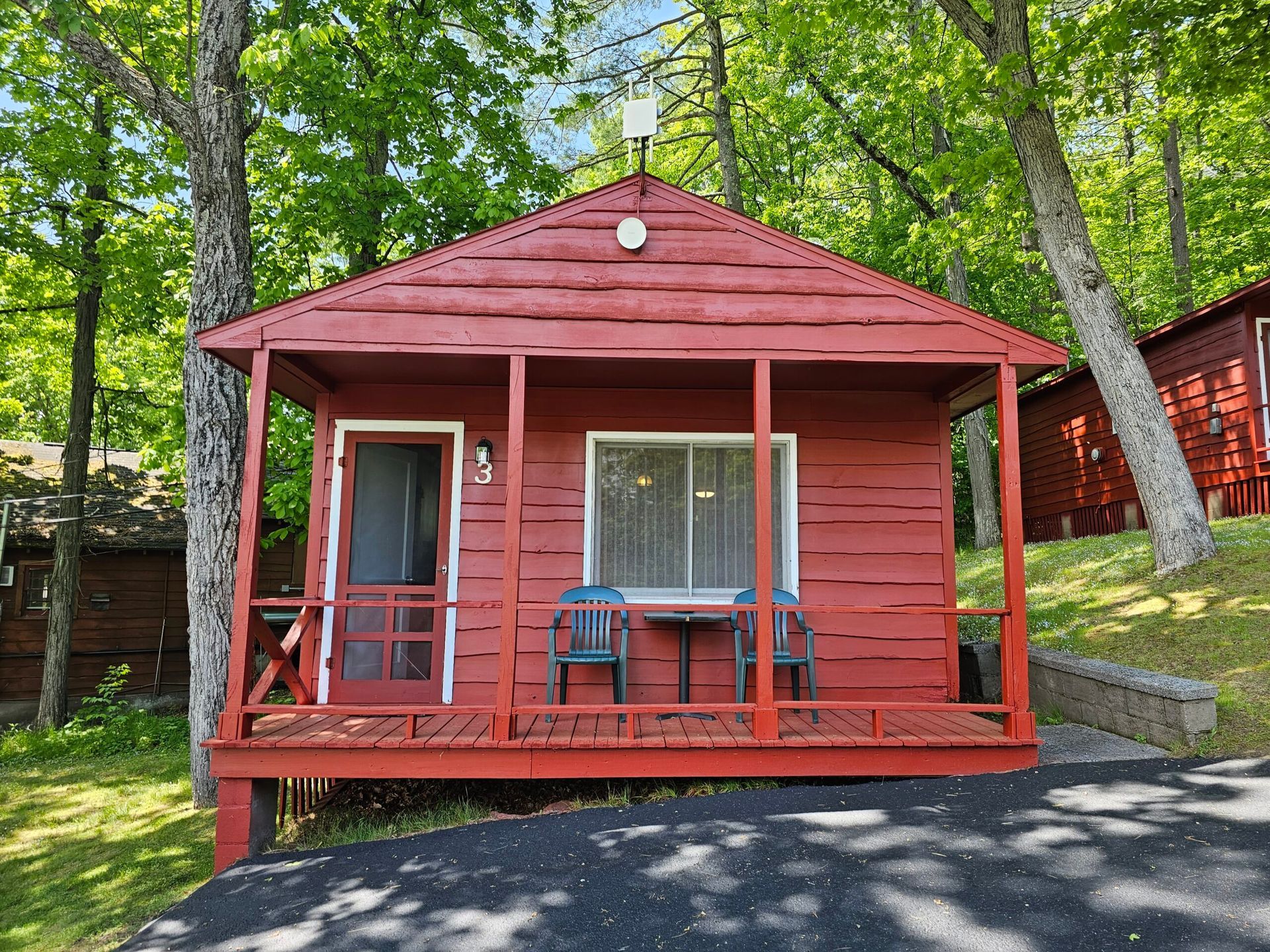 Small red cabin with porch, white door, and picnic table in a wooded area
