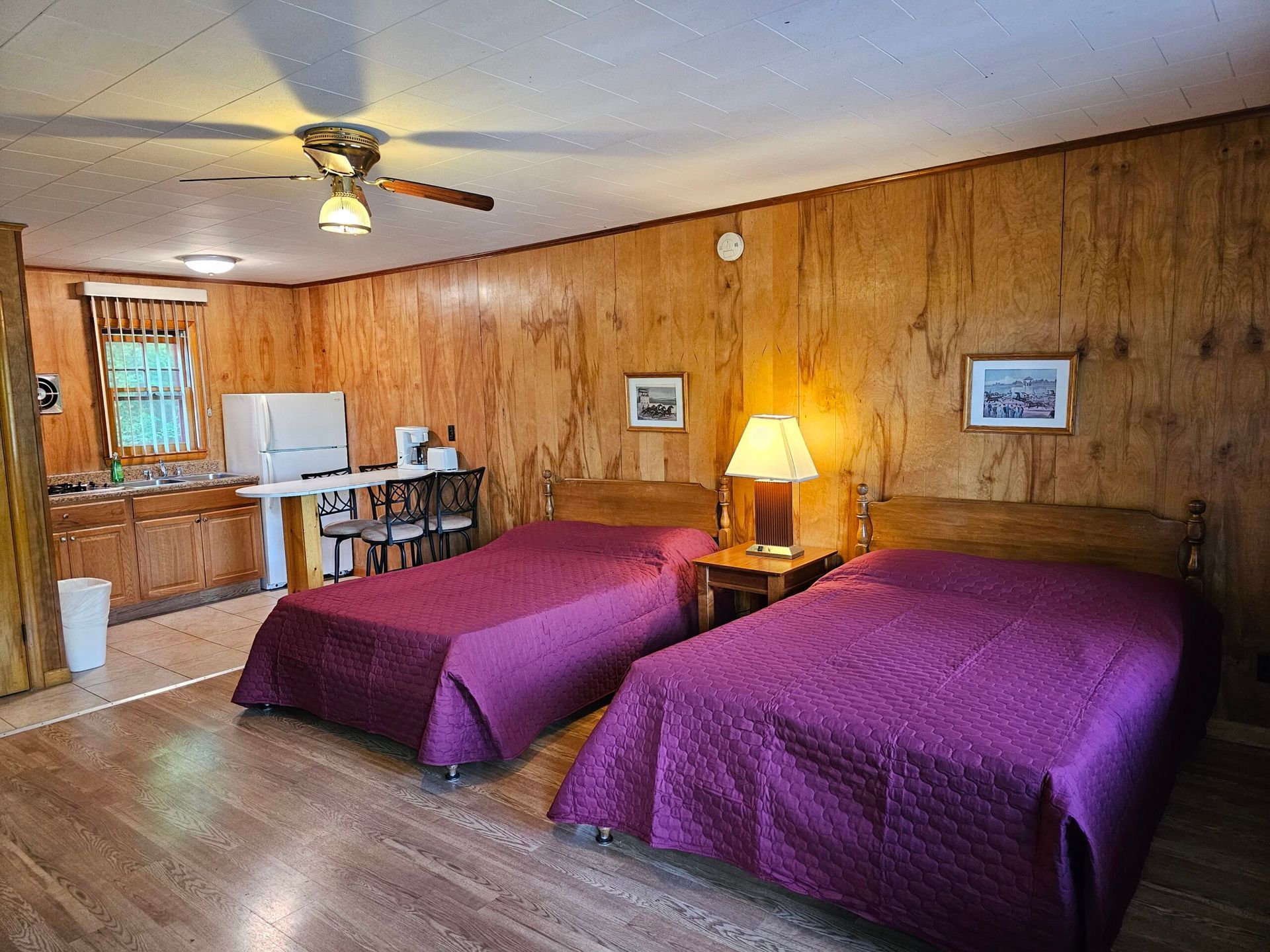 Wood-paneled motel room with two purple-covered beds, a lamp, ceiling fan, and small dining area