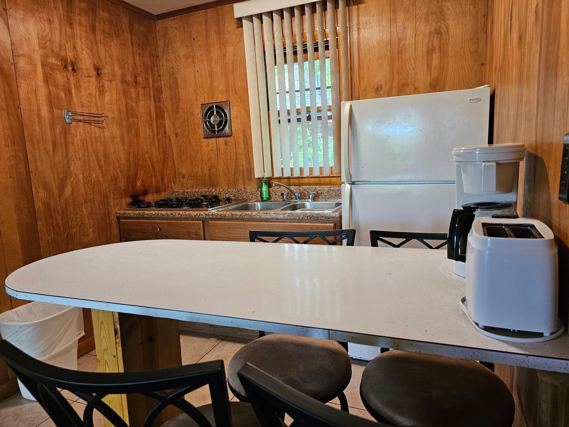 Small kitchen with a white table, black stools, wood-paneled walls, sink, and refrigerator.