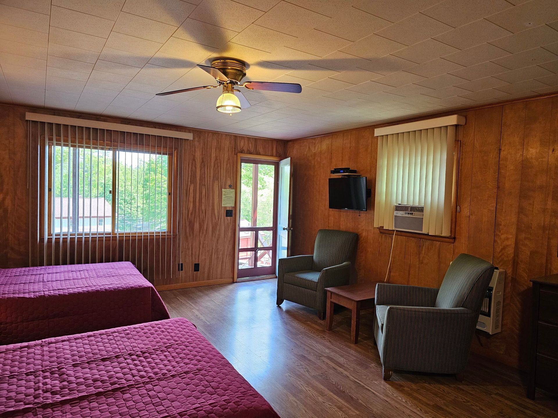 Wood-paneled bedroom with two twin beds, two chairs, a ceiling fan, and a sliding glass door.