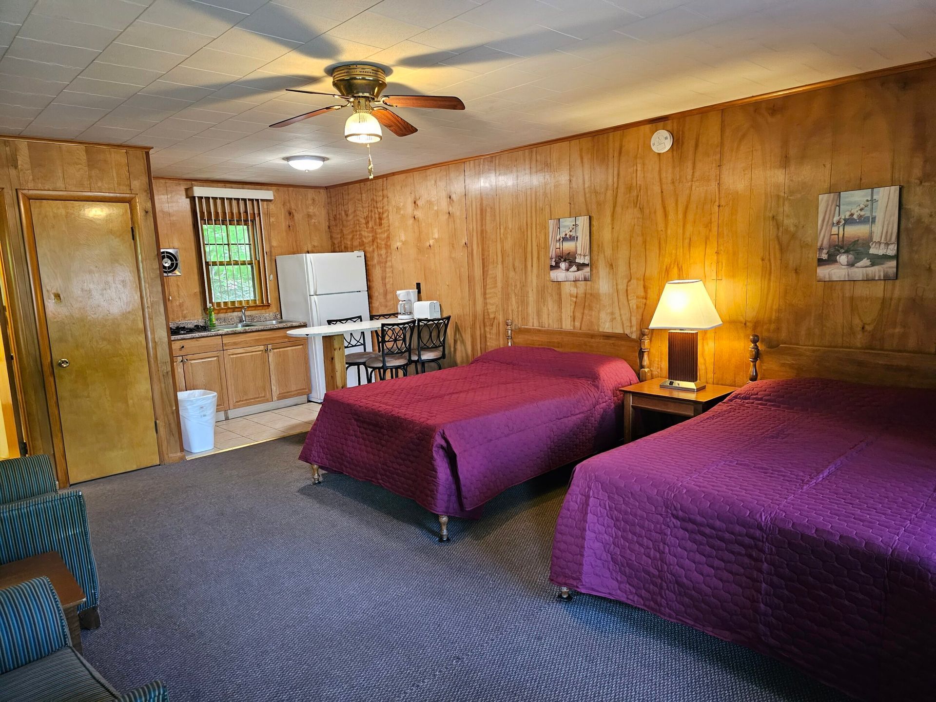Wood-paneled motel room with two purple beds, lamp, ceiling fan, and kitchenette.