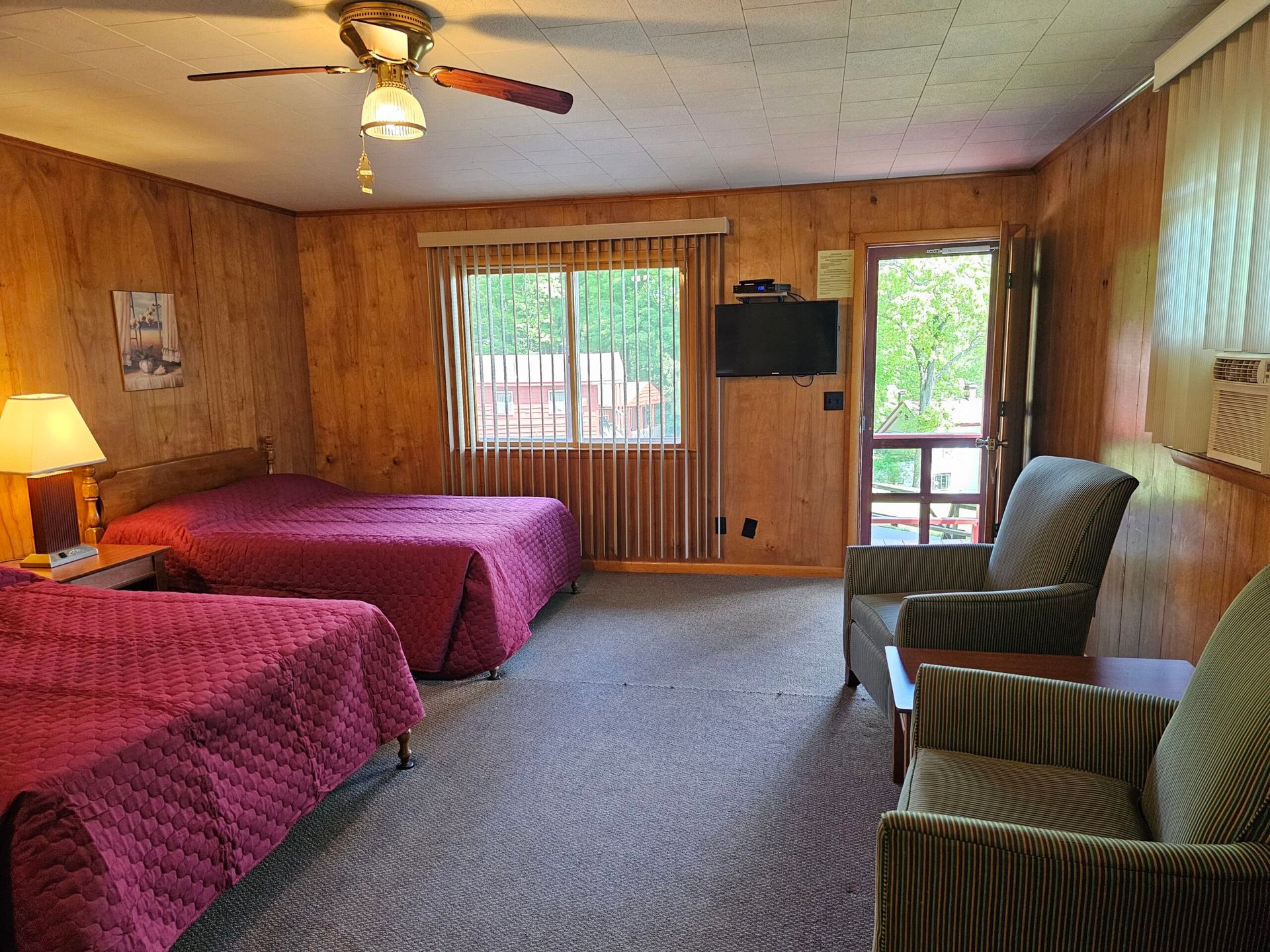 Wood-paneled bedroom with two beds, chairs, a window, and a ceiling fan.