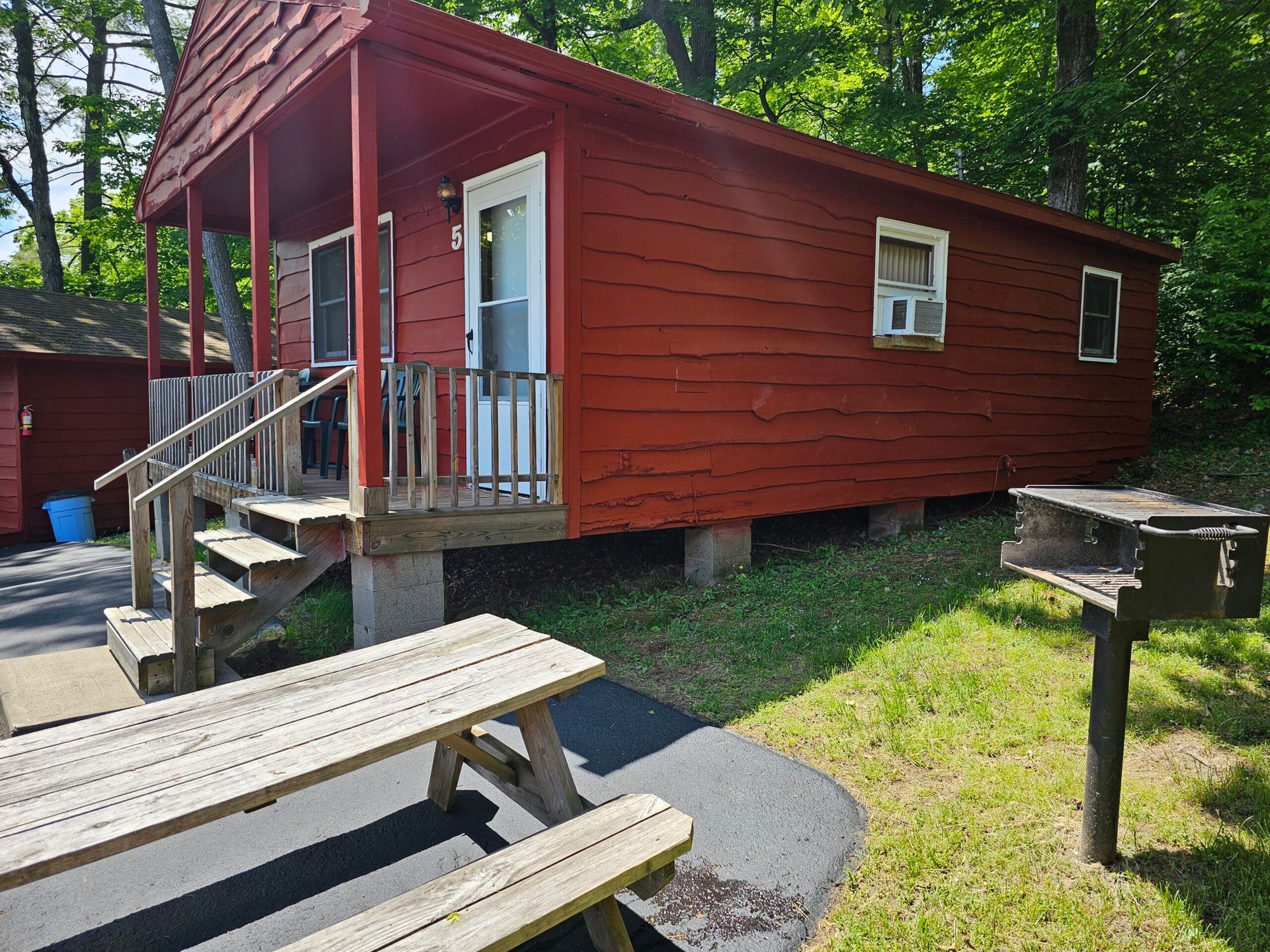 Red cabin with a small porch and steps in a wooded campsite, with picnic tables in front