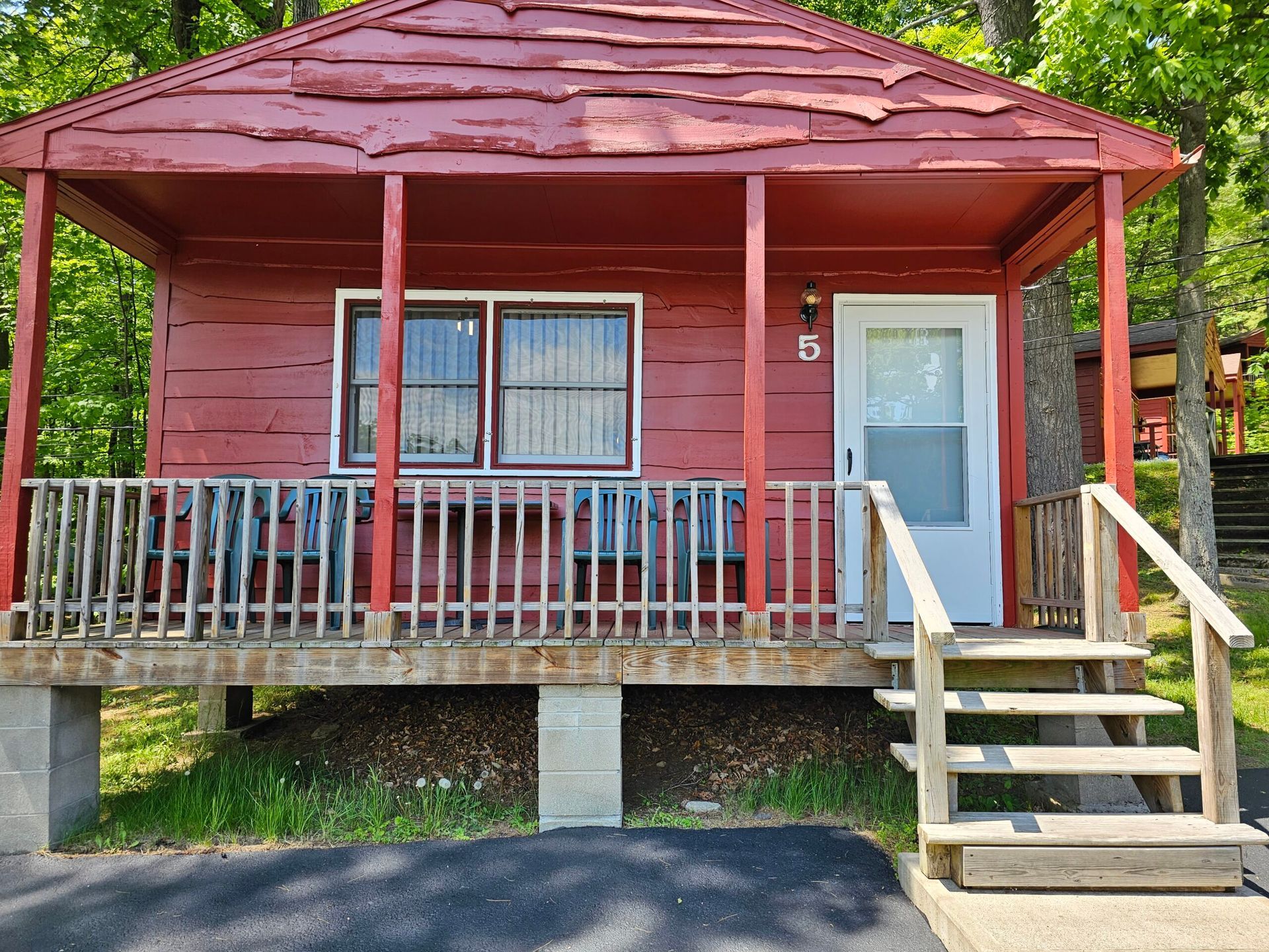 Red cabin porch with stairs, railing, and white door, set among trees.