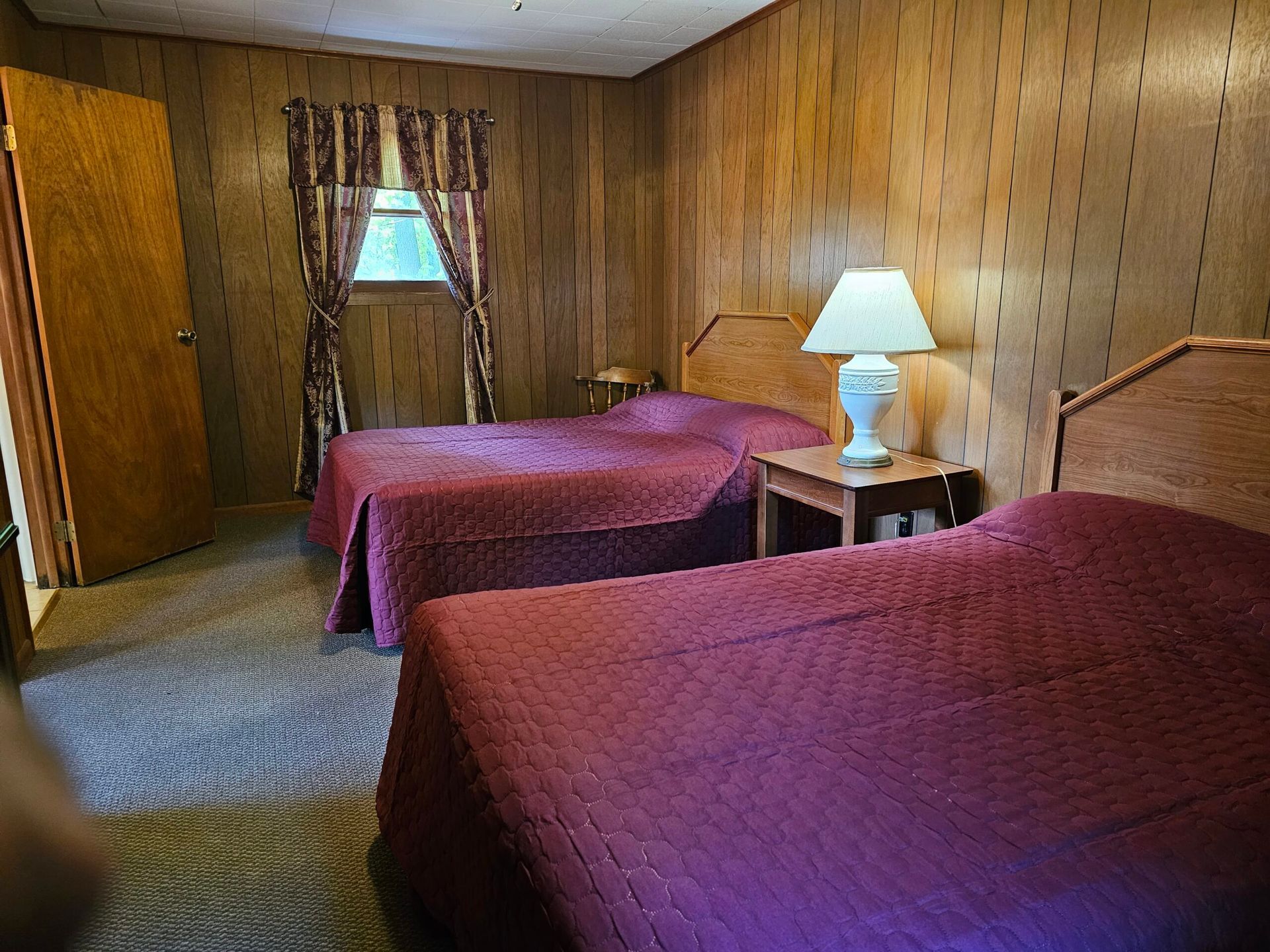 Small motel room with two beds, wood-paneled walls, a window, and a lamp on a bedside table.