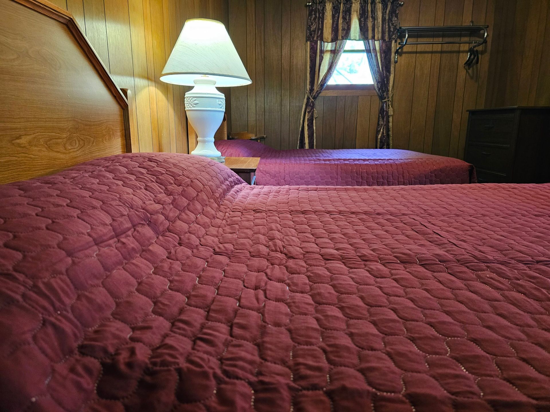 Two red quilted beds in a wood-paneled hotel room with a bedside lamp on.