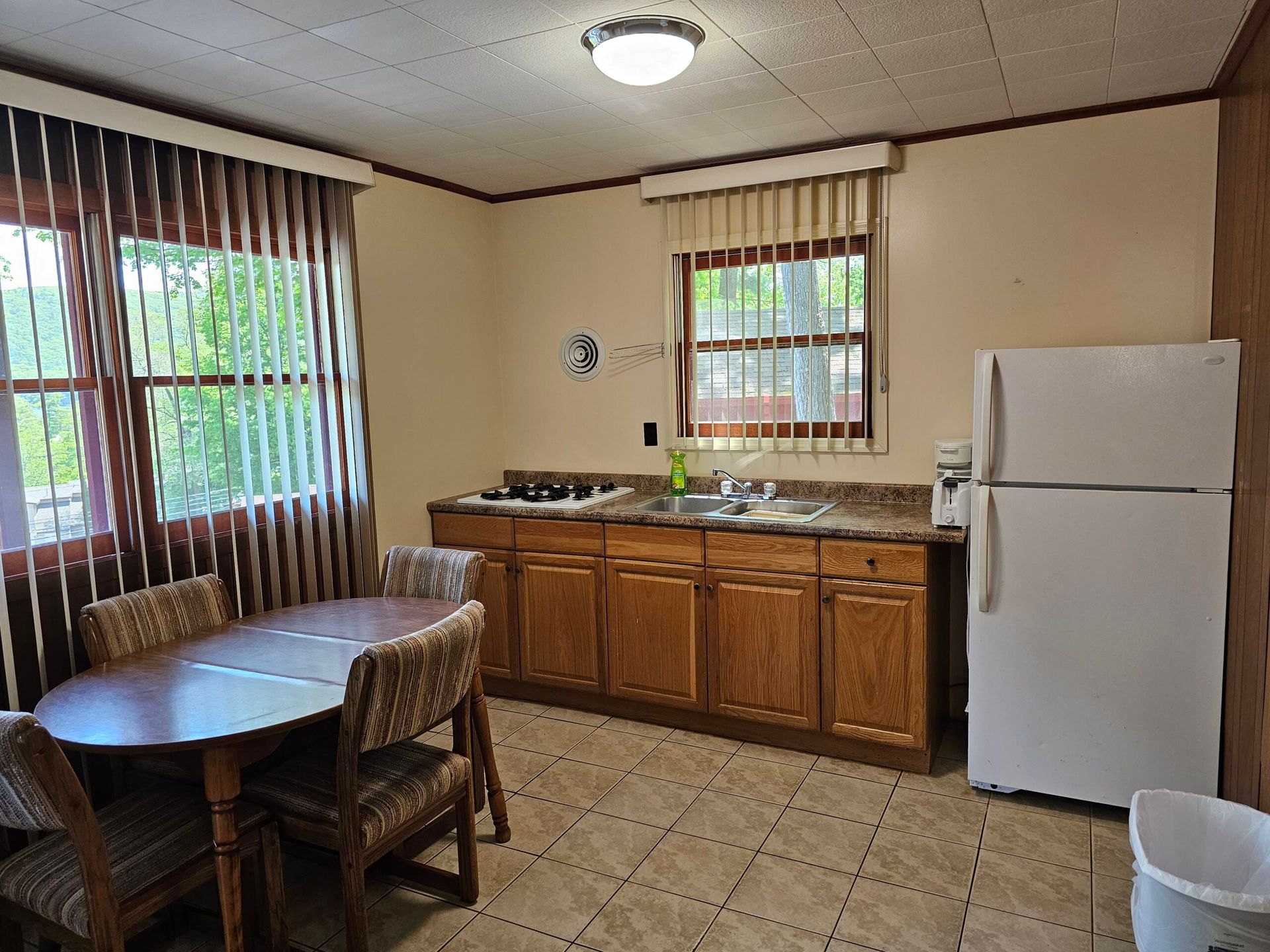 Small kitchen with wooden cabinets, white fridge, tiled floor, and a round table by the window.