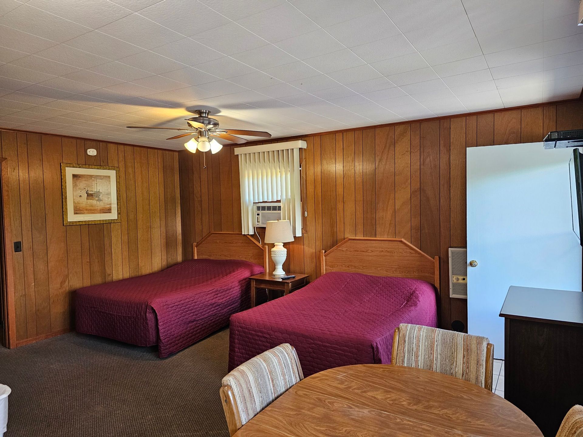 Wood-paneled motel room with two beds, ceiling fan, TV, and a small table by the window.