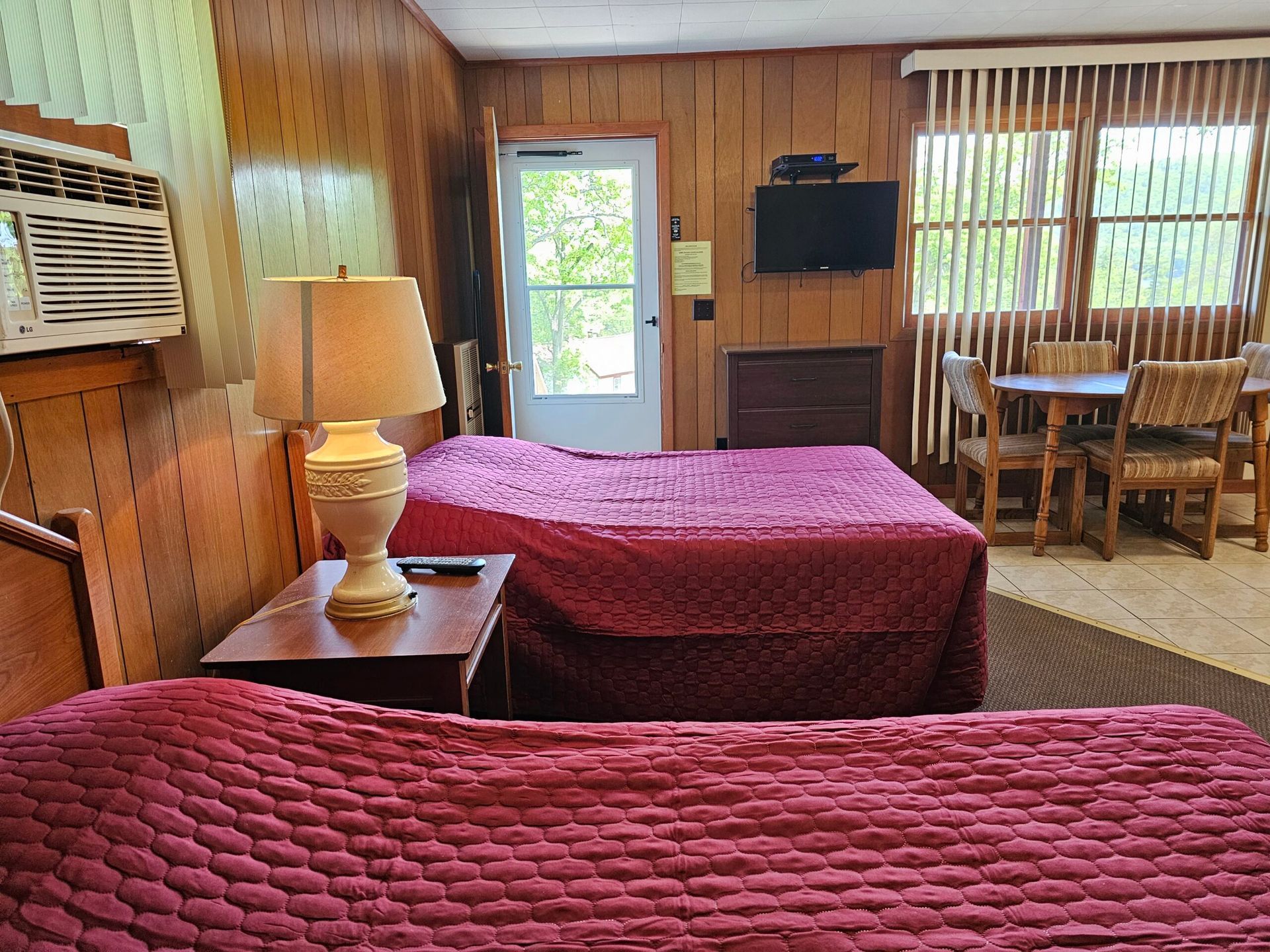 Wood-paneled motel room with two red beds, lamp, TV, and small table by a window