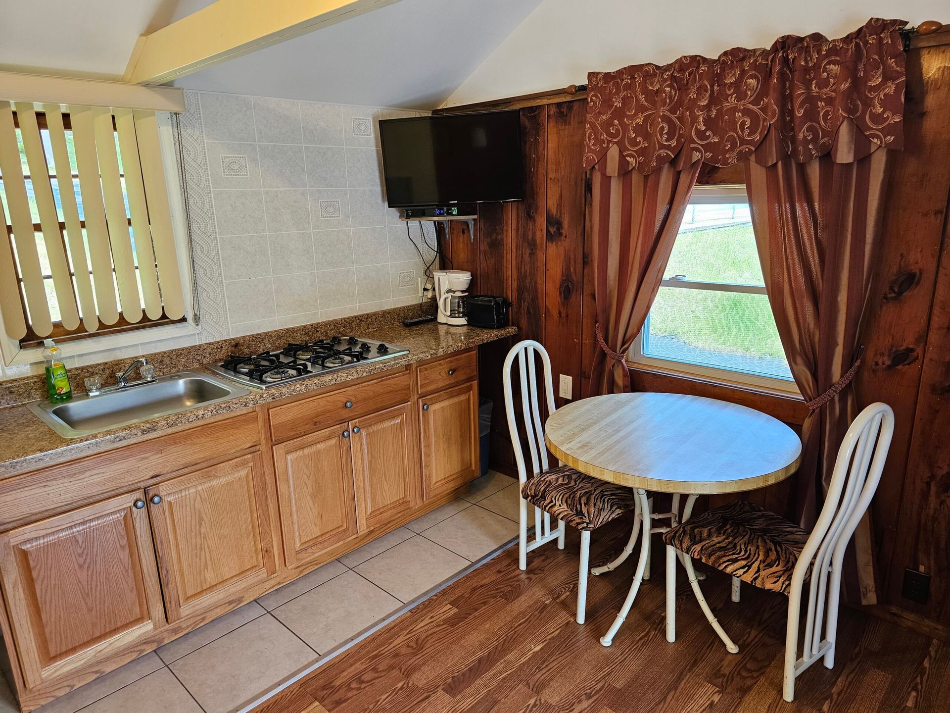 Small kitchen with wooden cabinets, sink, round table, and chairs by a curtained window