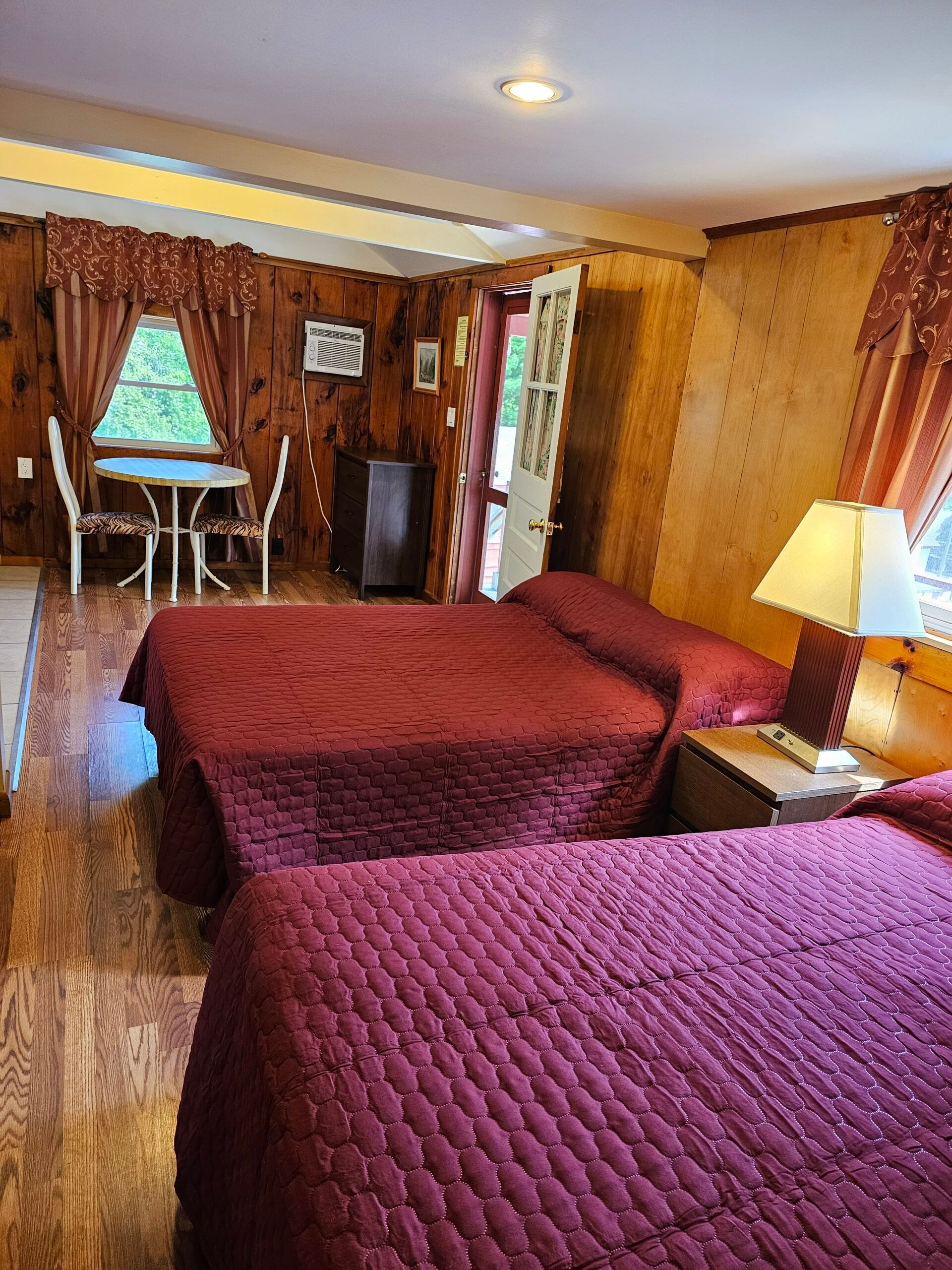 Wood-paneled motel room with two maroon beds, a lamp, and a small table by the window.
