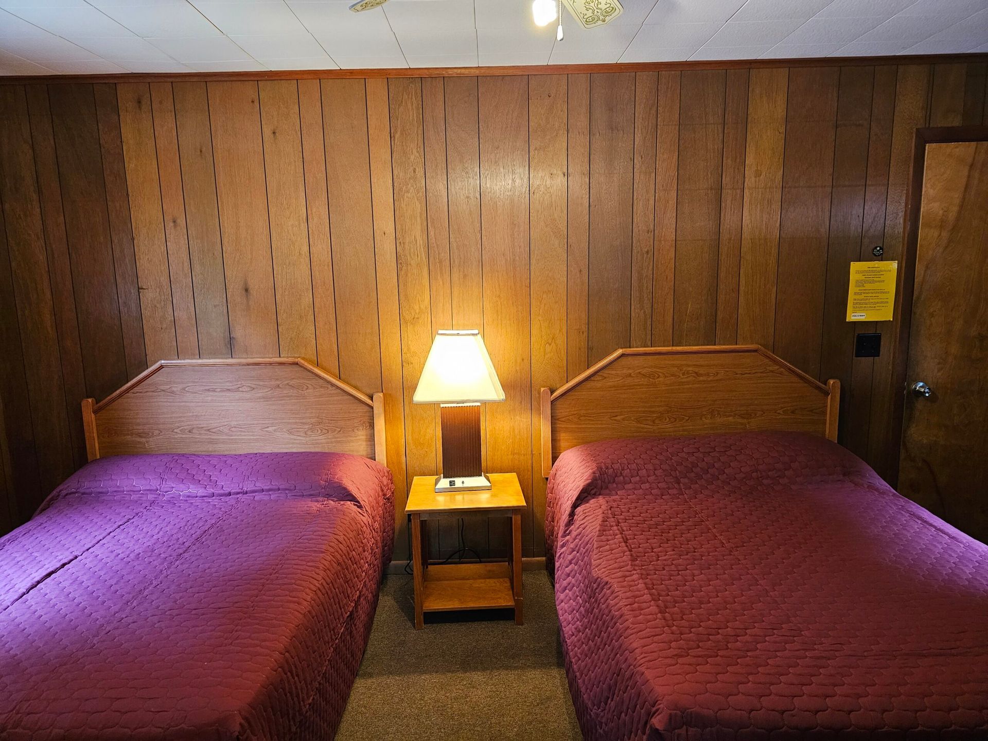 Two twin beds with red and purple bedspreads in a wood-paneled motel room, with a lit lamp between them.