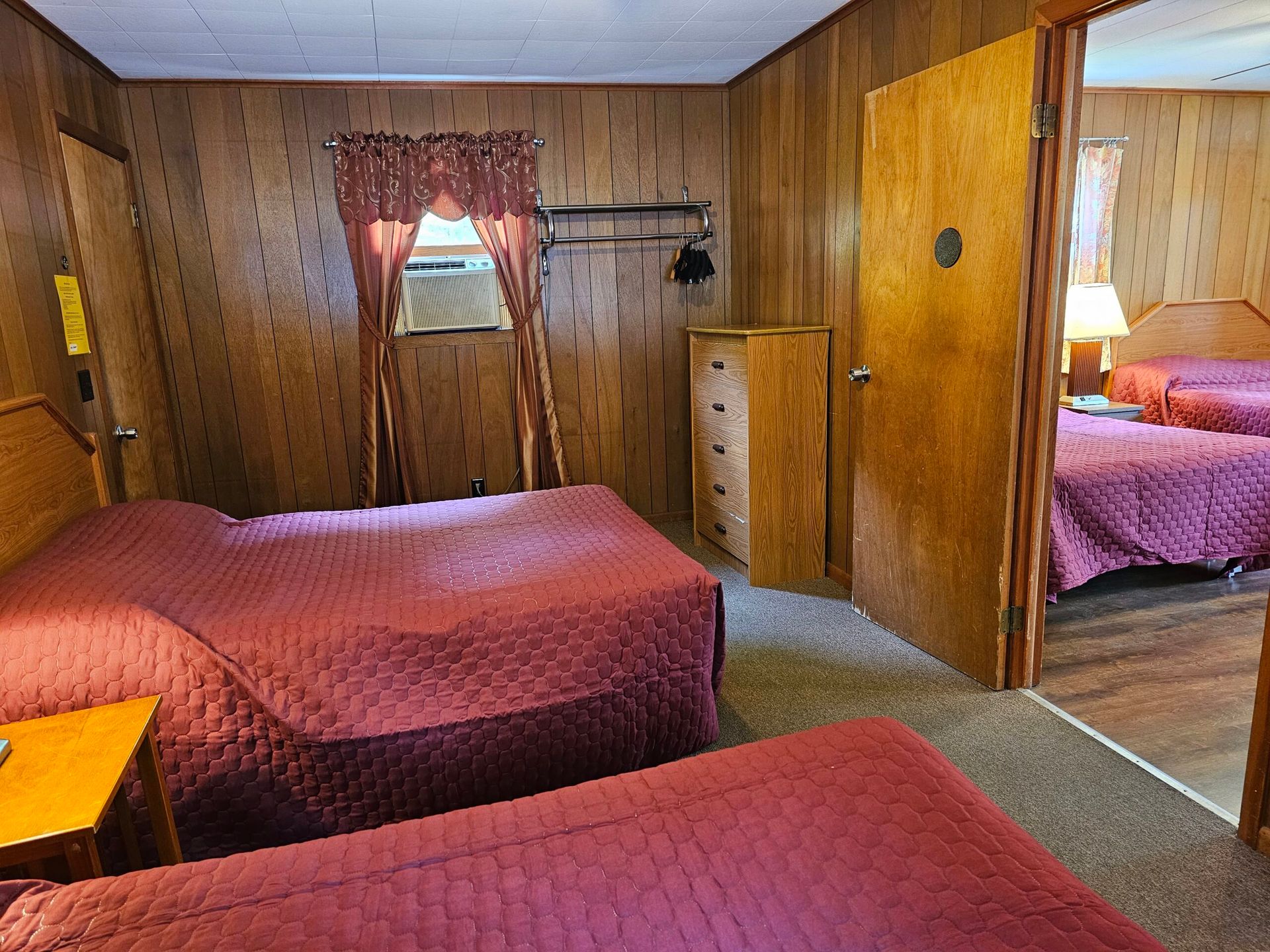 Wood-paneled bedroom with two beds, a small window, dresser, and an open doorway to a second bed.