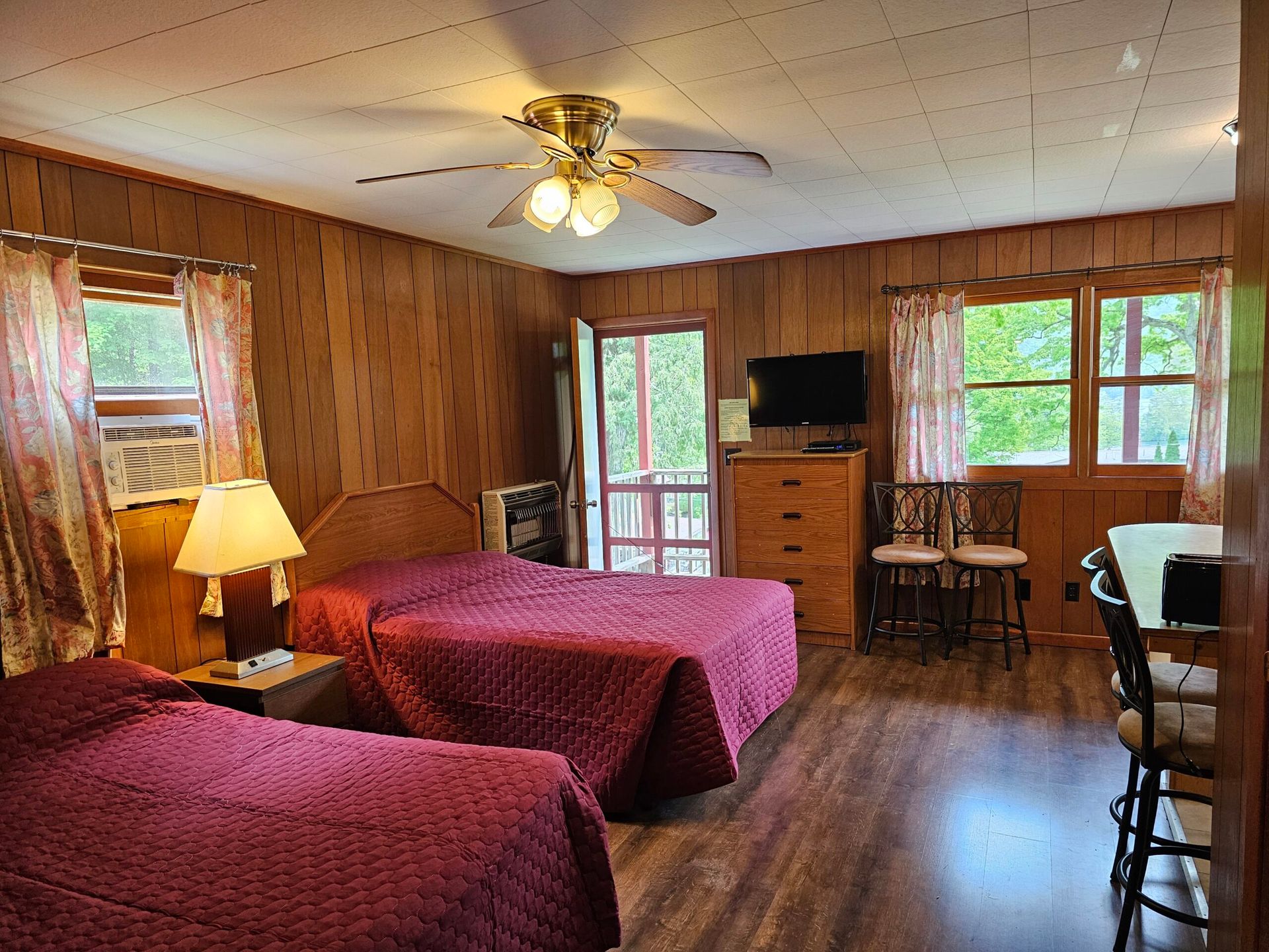 Wood-paneled motel room with two burgundy beds, TV, dresser, ceiling fan, and dining area by windows