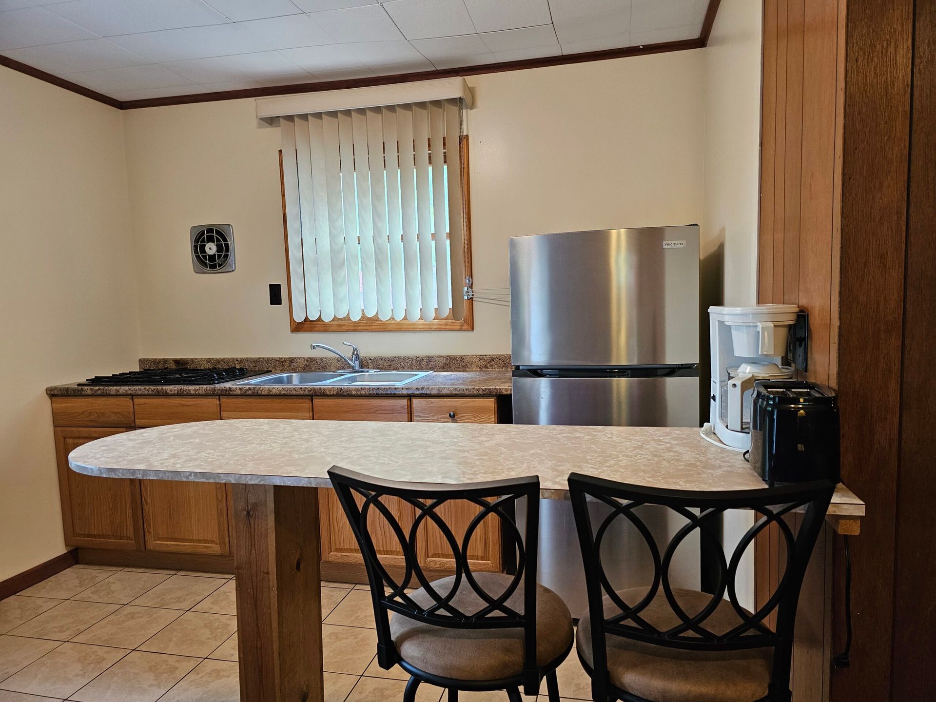 Small kitchen with wooden cabinets, white island, two black chairs, and stainless steel refrigerator.